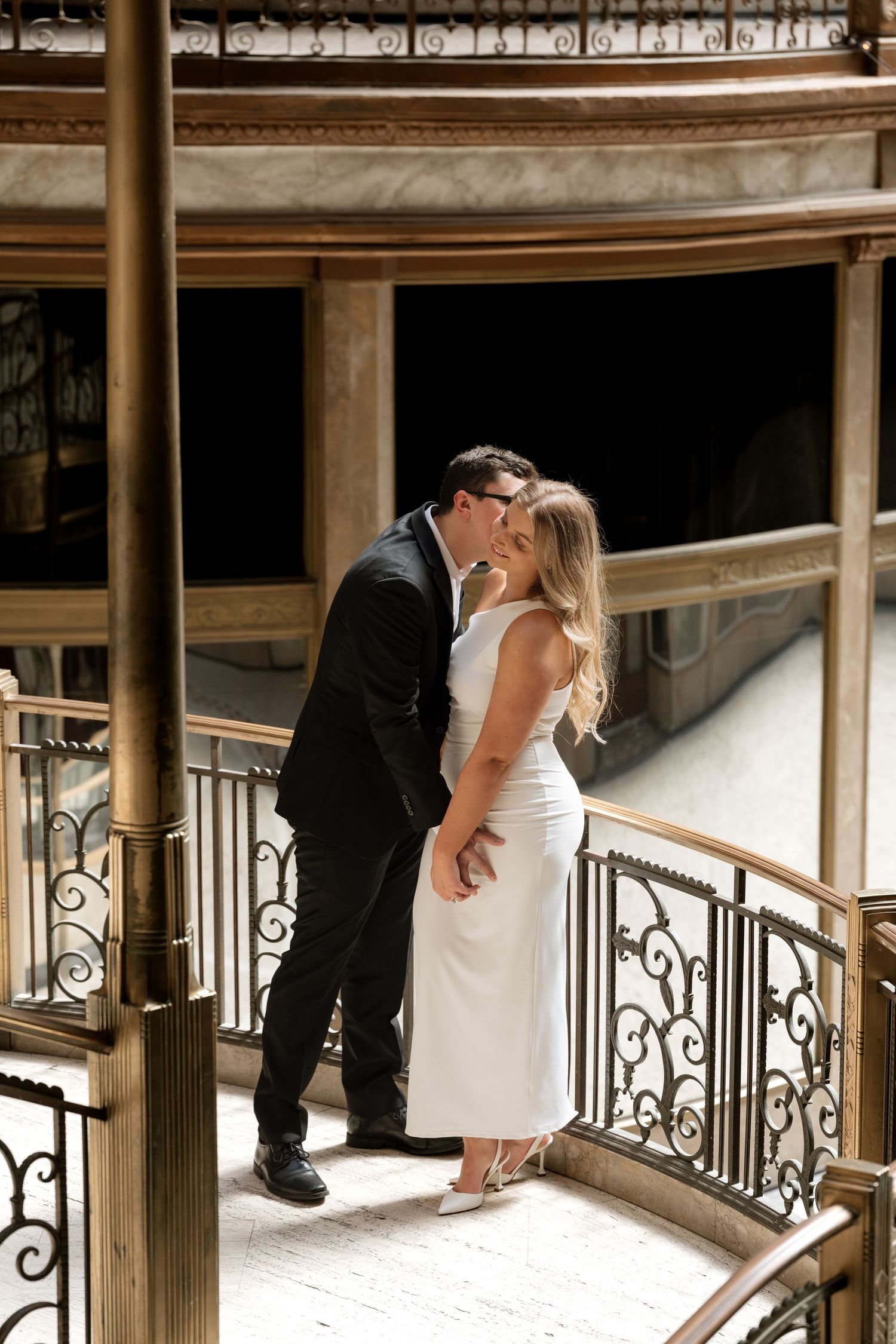 Couple sharing a romantic kiss on an ornate balcony staircase, woman in white dress, man in black suit.
