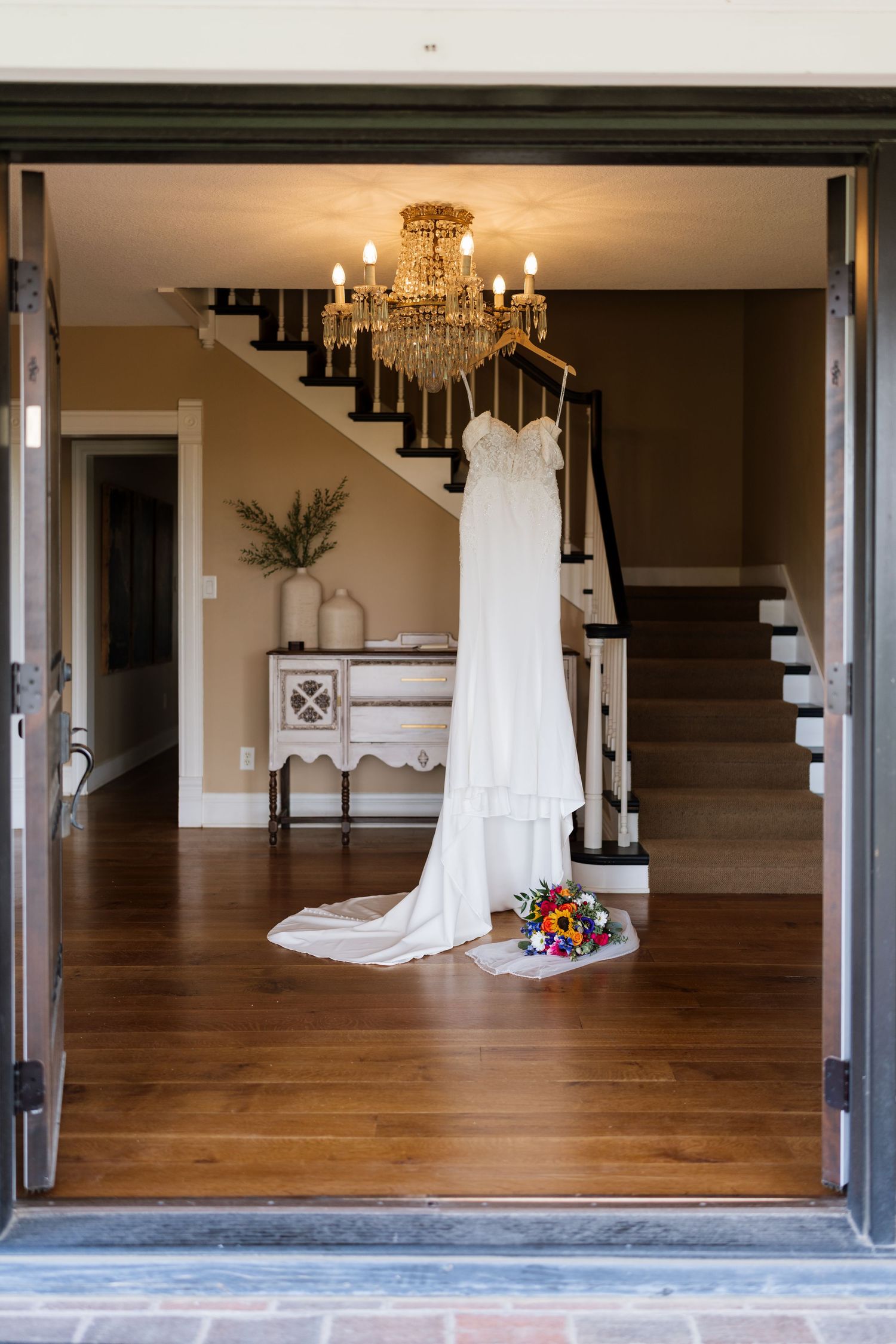 White wedding dress hanging from chandelier in elegant foyer with staircase and bridal bouquet on hardwood floor.