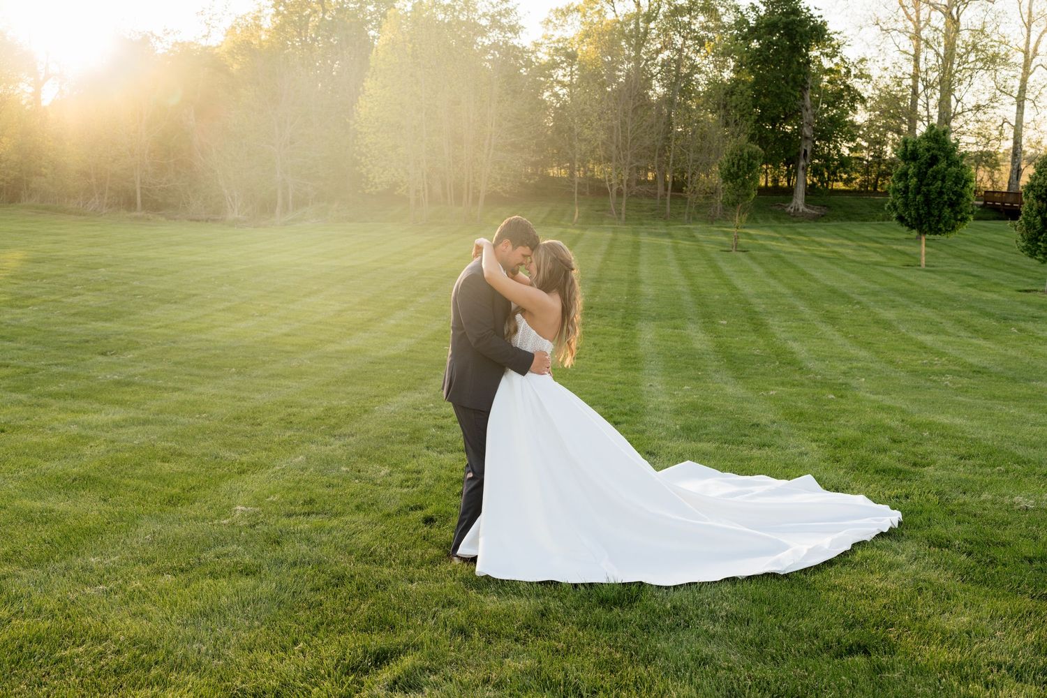 Bride and groom sharing a romantic kiss at golden hour on a lush green lawn with sun rays glowing behind them.