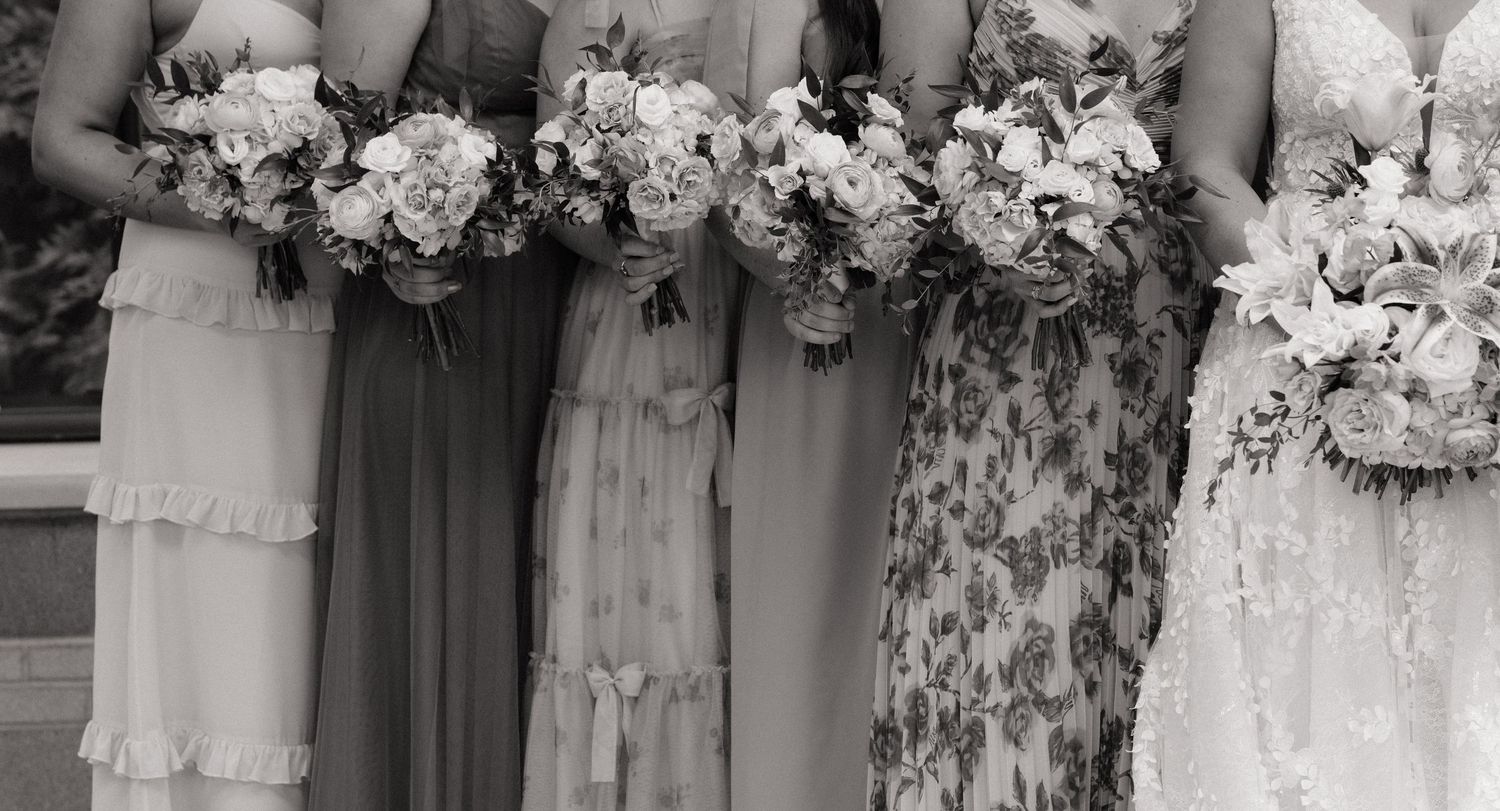 Black and white photo of bridesmaids in mismatched dresses holding floral bouquets standing beside a bride in a white gown.