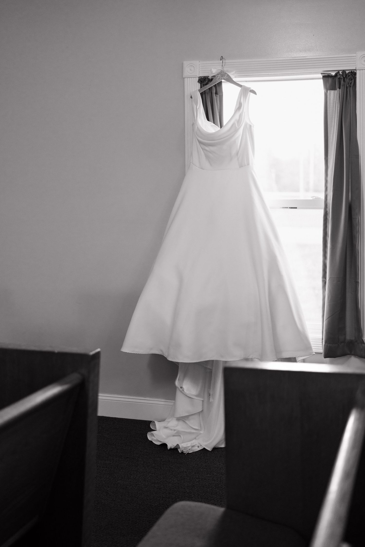 Classic white wedding gown hanging in window with dramatic lighting and dark interior.