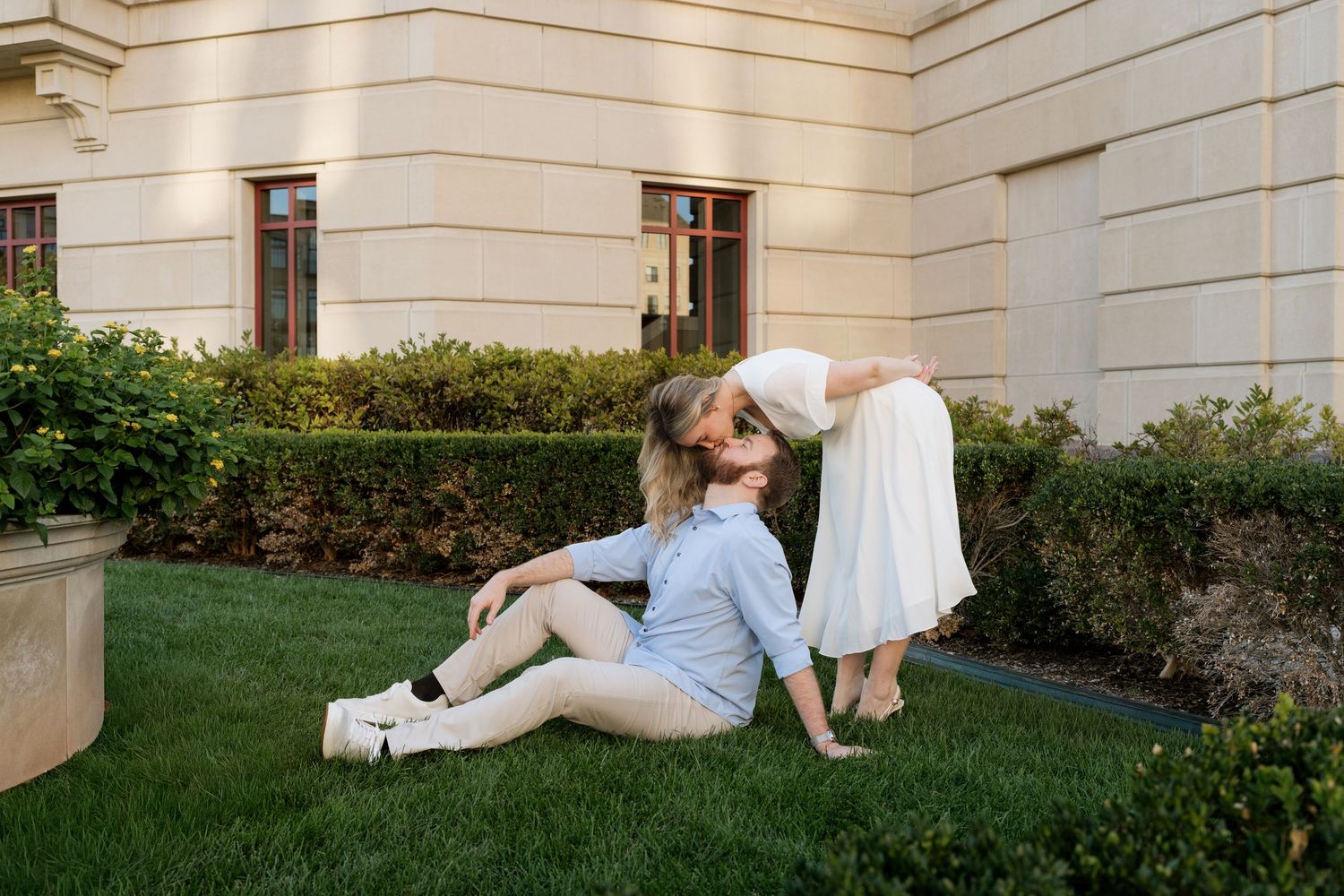 Couple sharing a romantic kiss on green lawn outside elegant building, woman in white dress, man in light blue outfit.