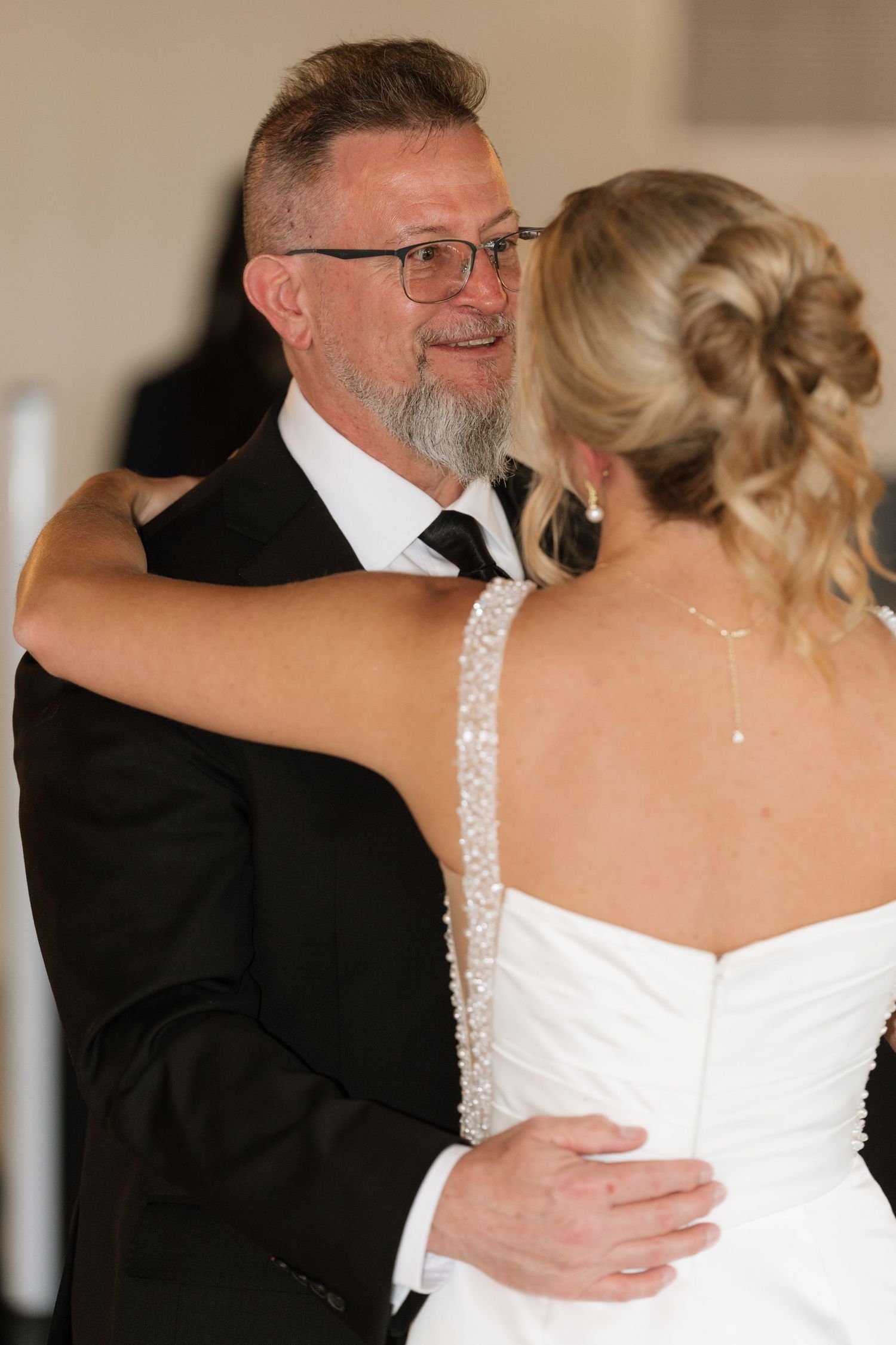 A bearded man in a black suit shares a tender dance with a bride in a white beaded gown at a wedding reception.