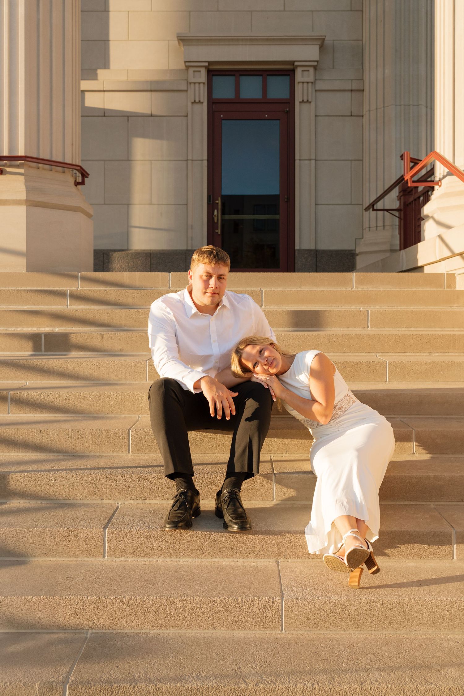 Bride in white dress rests head on groom's shoulder as they sit together on church steps in golden hour light.