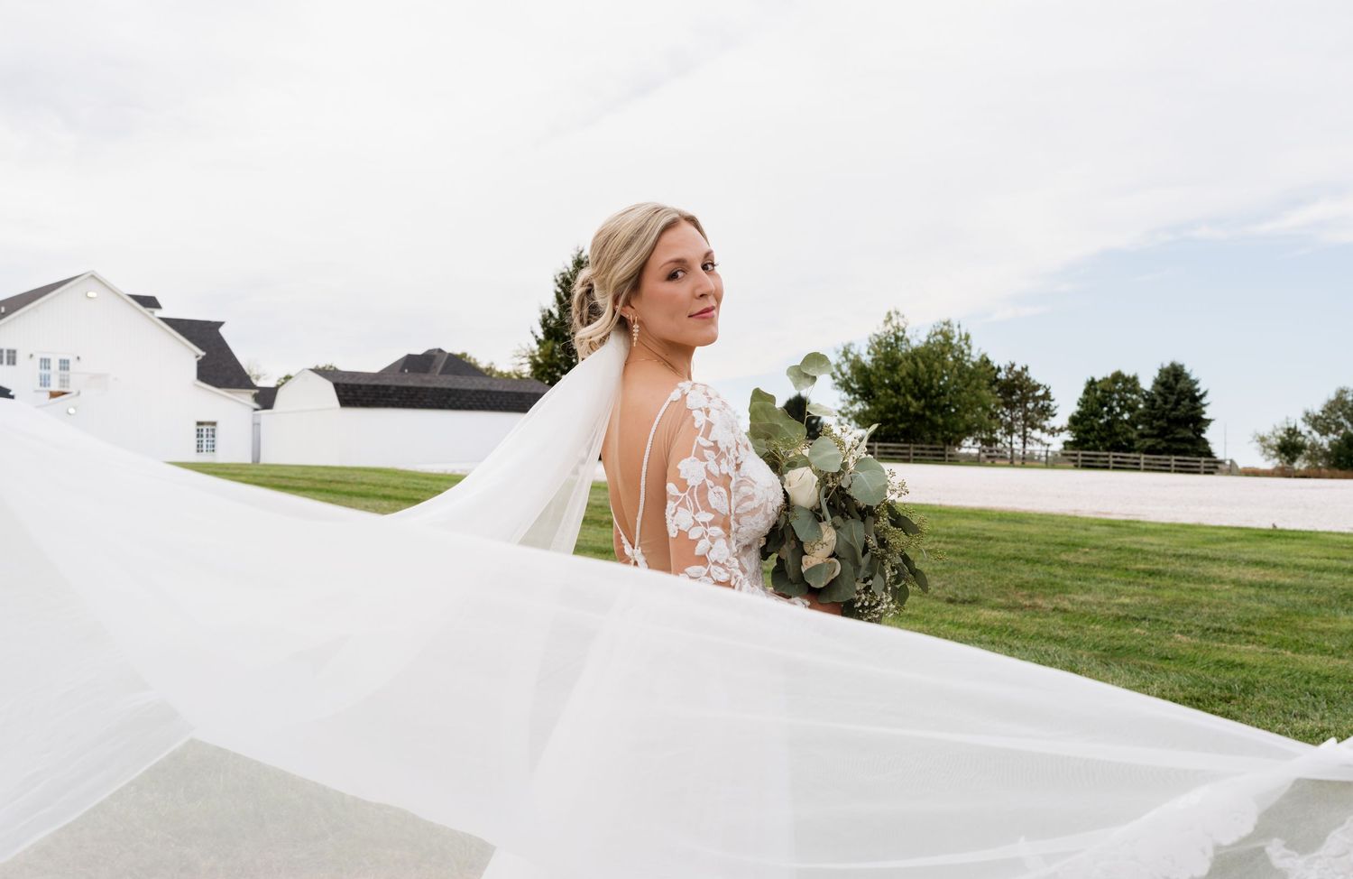 Bride in a lace wedding dress with flowing veil holds a greenery bouquet outdoors near a white barn.