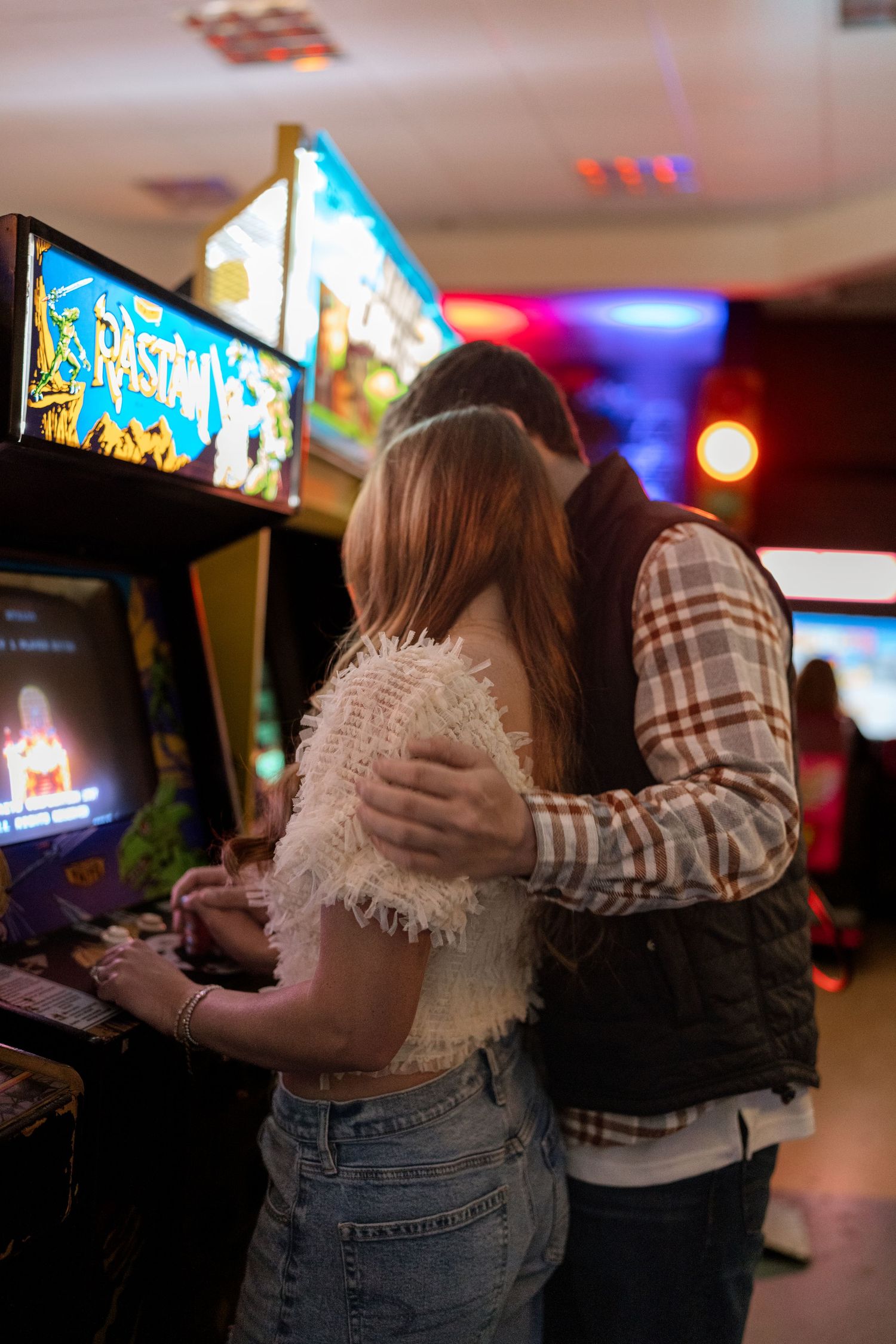 Two people in a dimly lit arcade embrace while playing video games in a romantic moment.