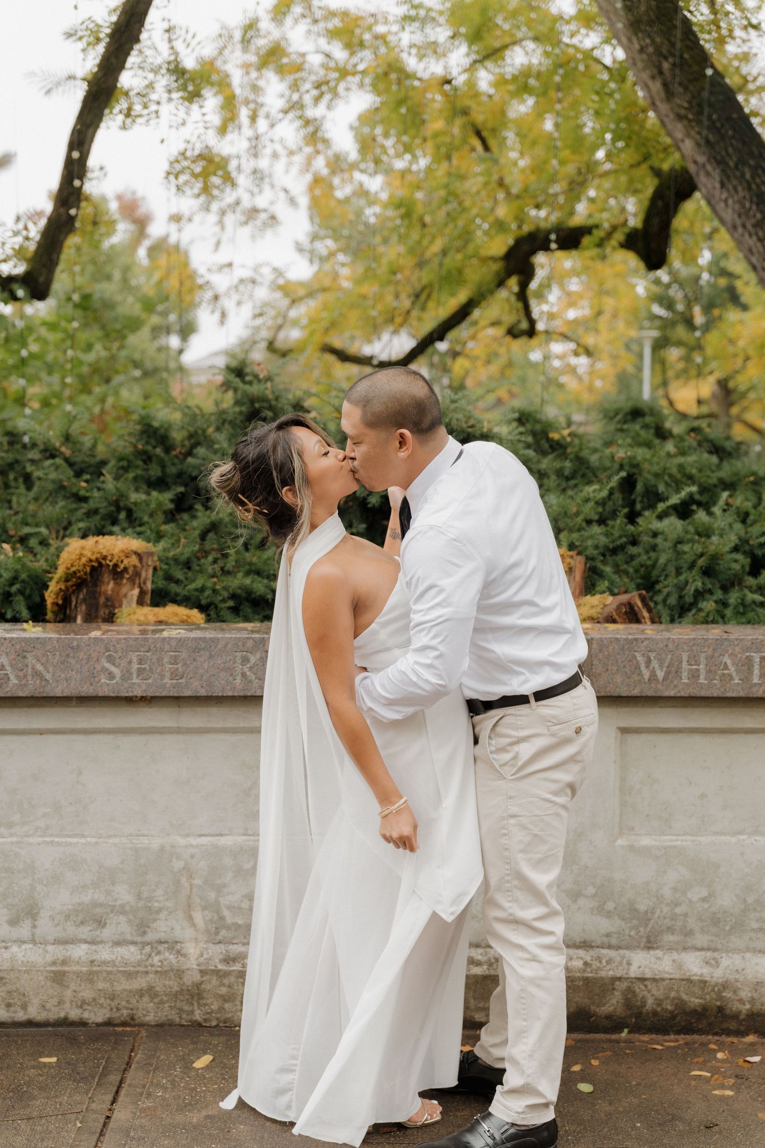 A couple in white attire share romantic moments against a stone wall backdrop during an autumn outdoor photoshoot.