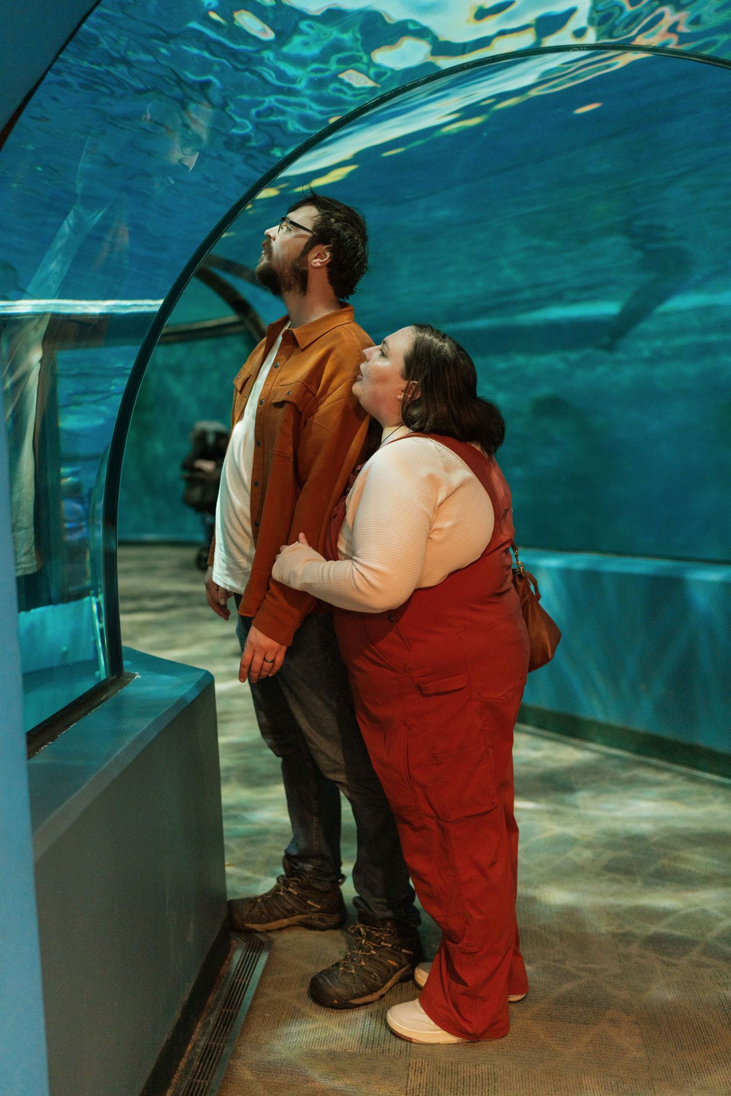 Two visitors observe marine life through a curved underwater viewing tunnel at an aquarium with turquoise blue water above.