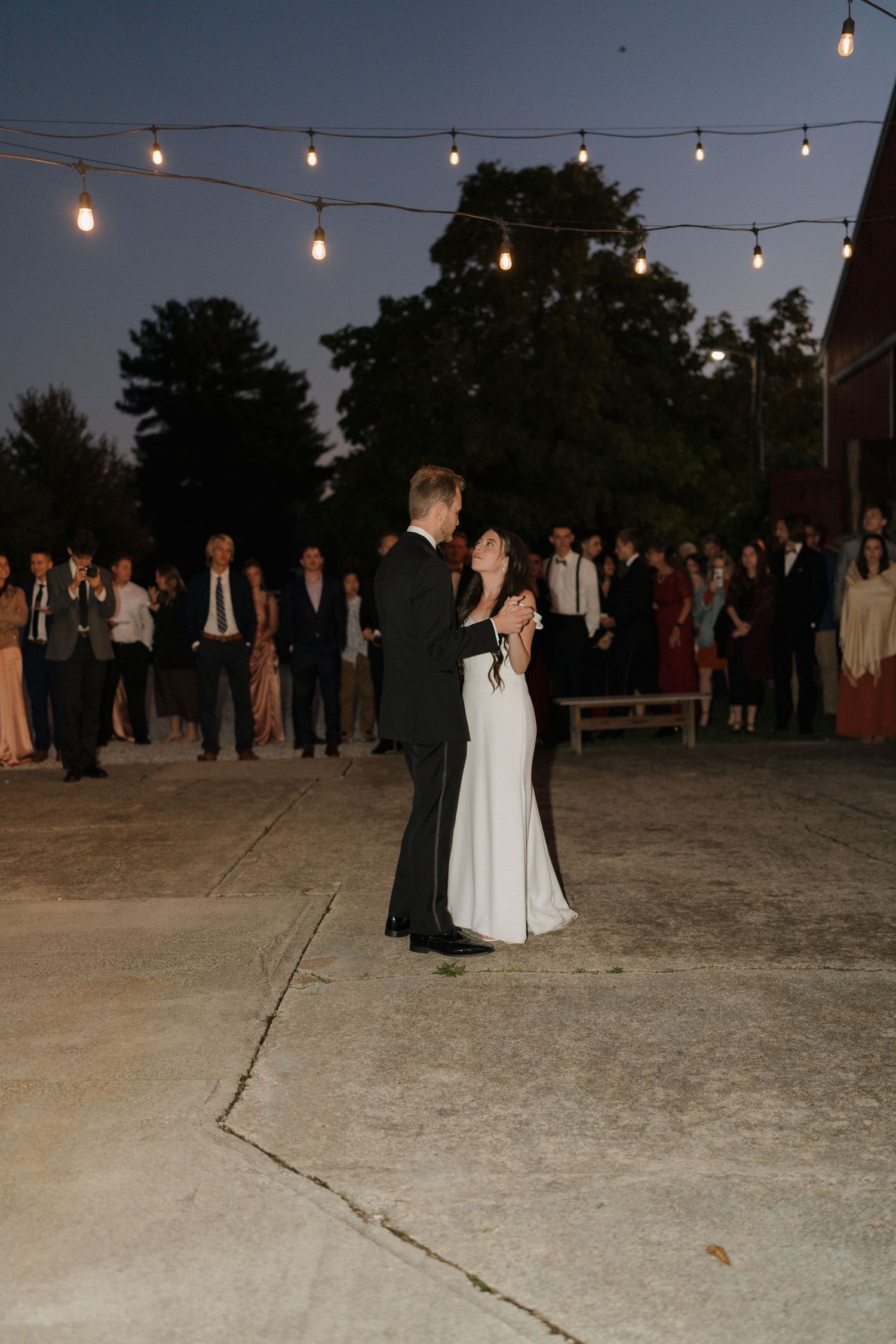 Wedding couple dances under string lights at outdoor evening reception with guests in background.