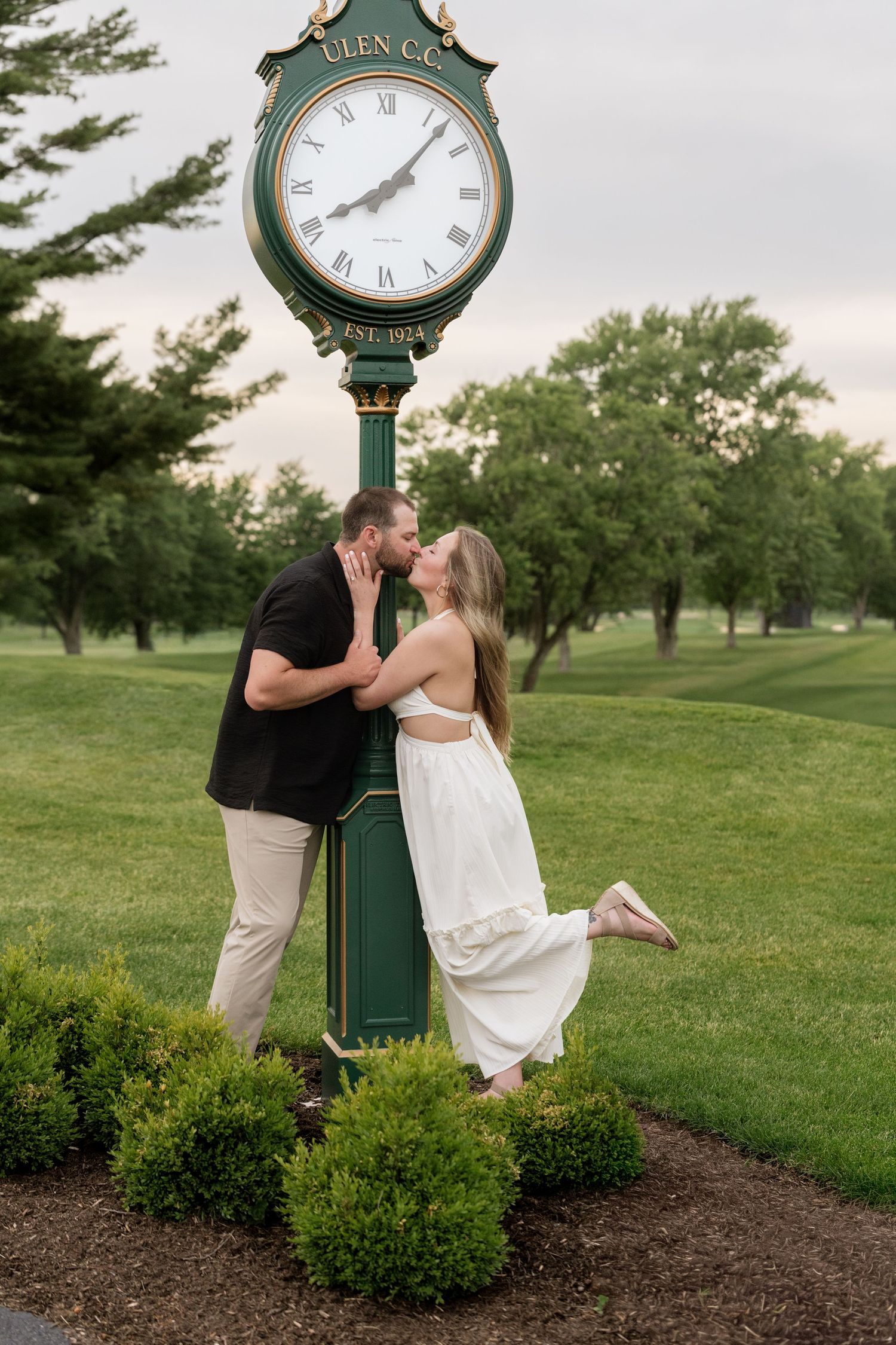 Couple kissing beside a green Ulen CC golf club clock tower surrounded by lush greenery during an engagement photo session.