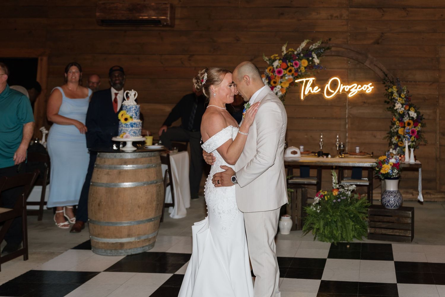 Bride and groom share first dance kiss at rustic wedding venue with checkered floor and neon sign.
