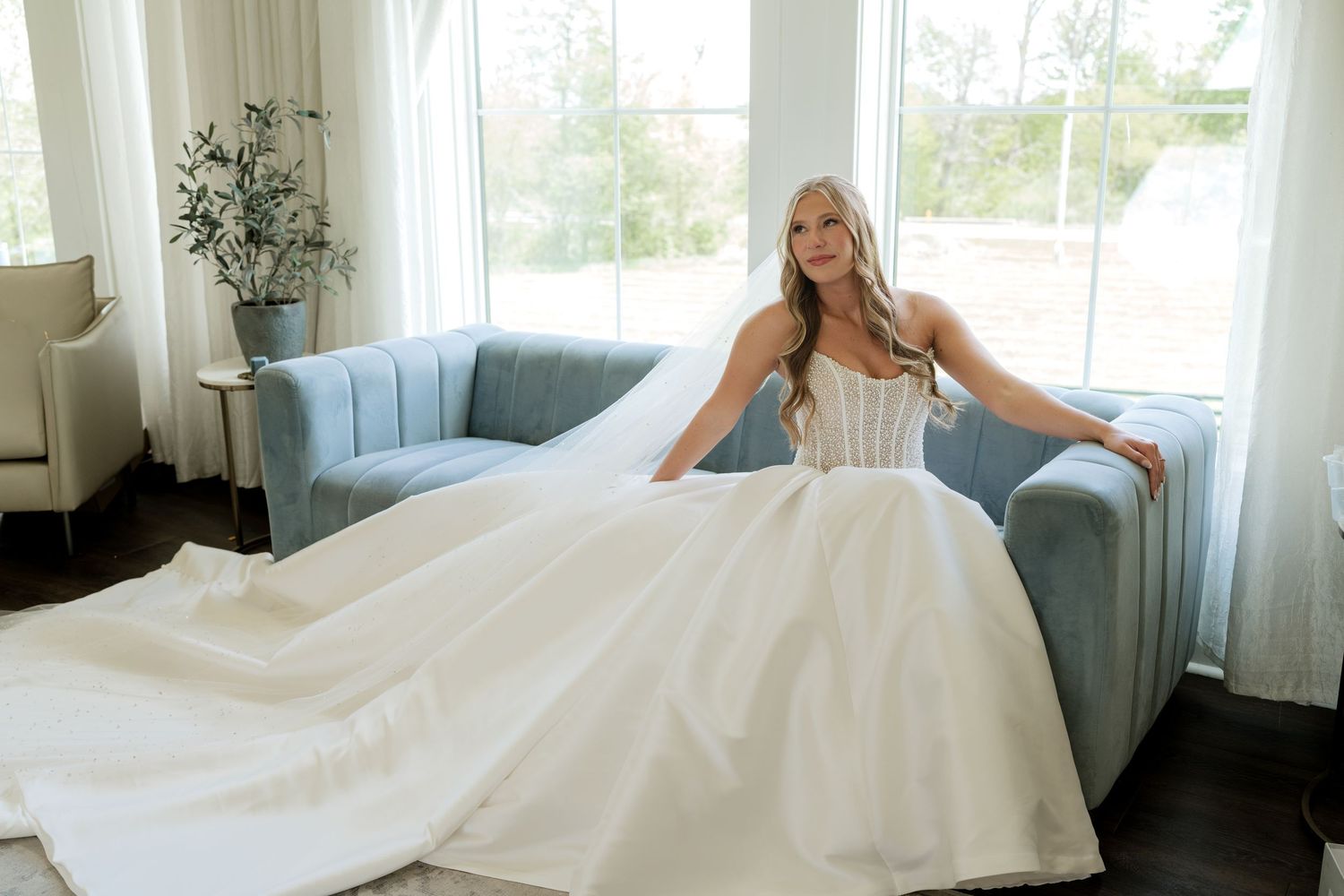 Bride in a stunning white ballgown wedding dress seated on a blue velvet sofa in an elegant bright room.