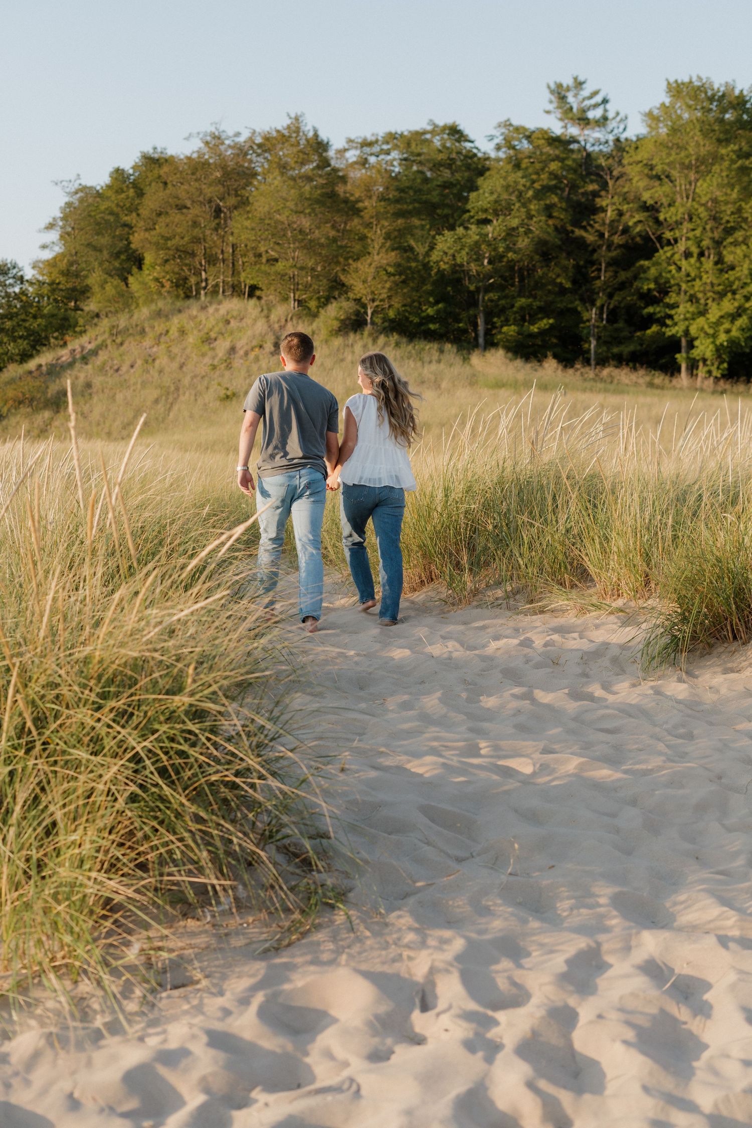 Couple walking hand in hand along sandy beach path with beach grass and trees in late afternoon light.