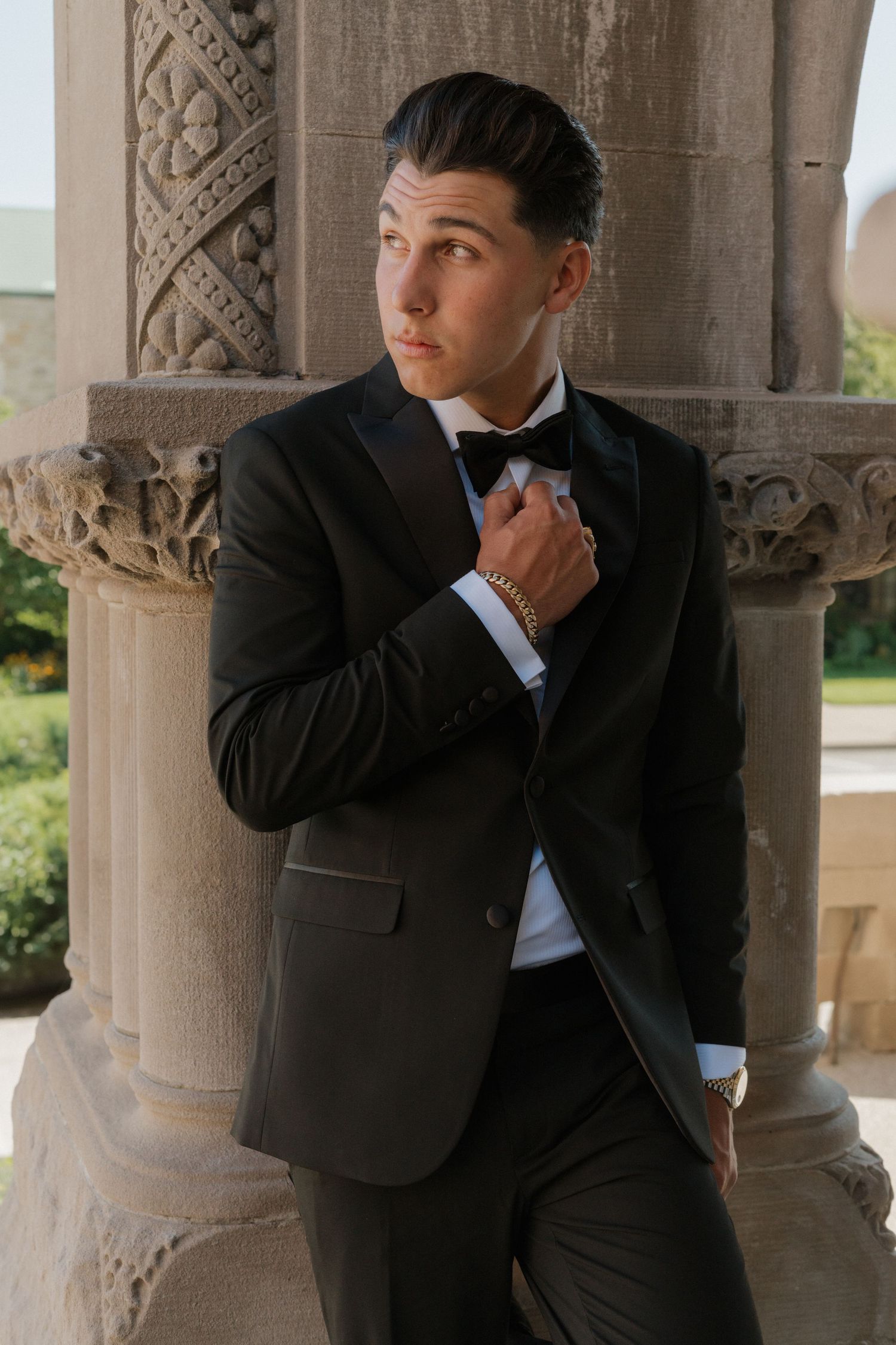 Groom in classic black tuxedo poses thoughtfully against stone columns.
