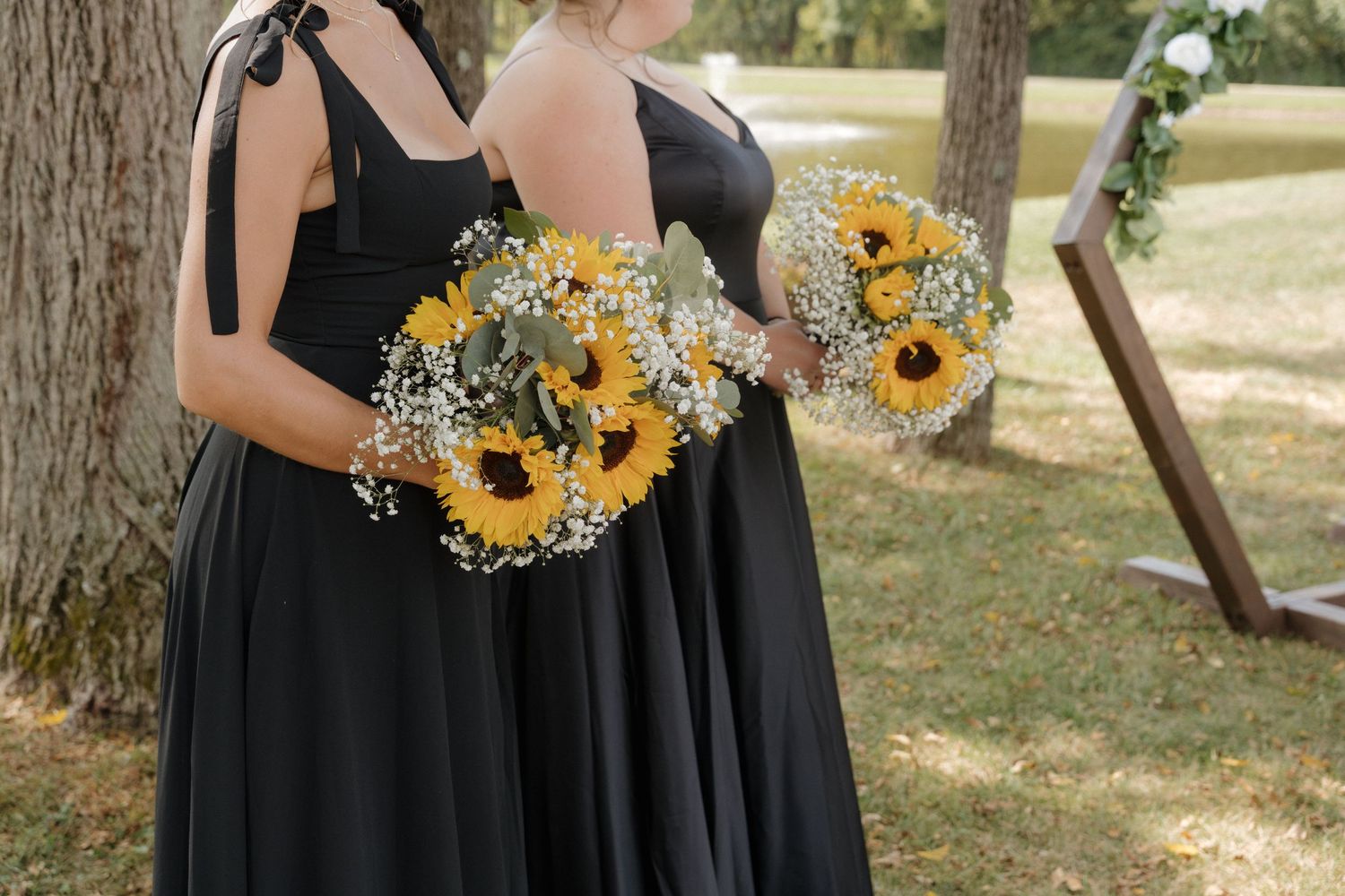 Wedding party holding sunflower bouquets against black dresses in outdoor setting.