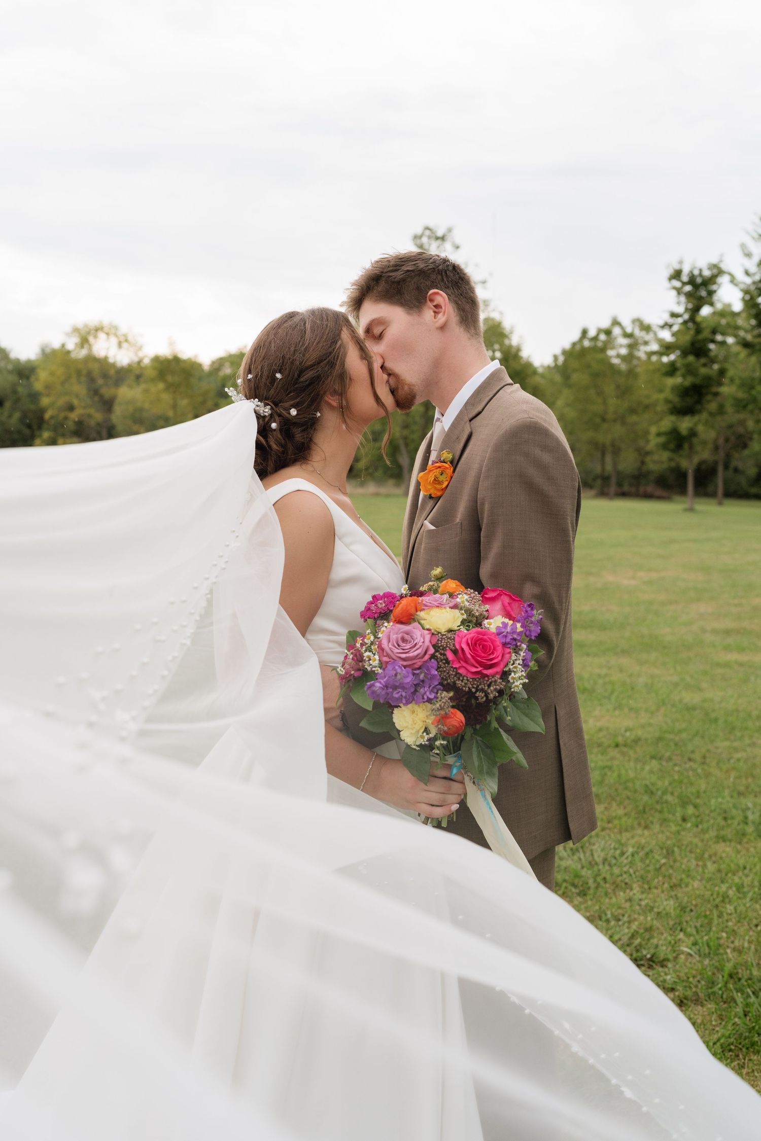 Bride and groom sharing a kiss outdoors, veil flowing in wind, holding colorful bridal bouquet.