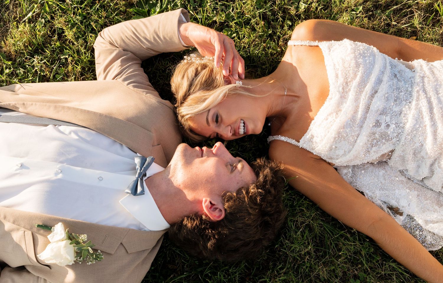 Bride and groom lying on green grass, smiling at each other in wedding attire, aerial view.