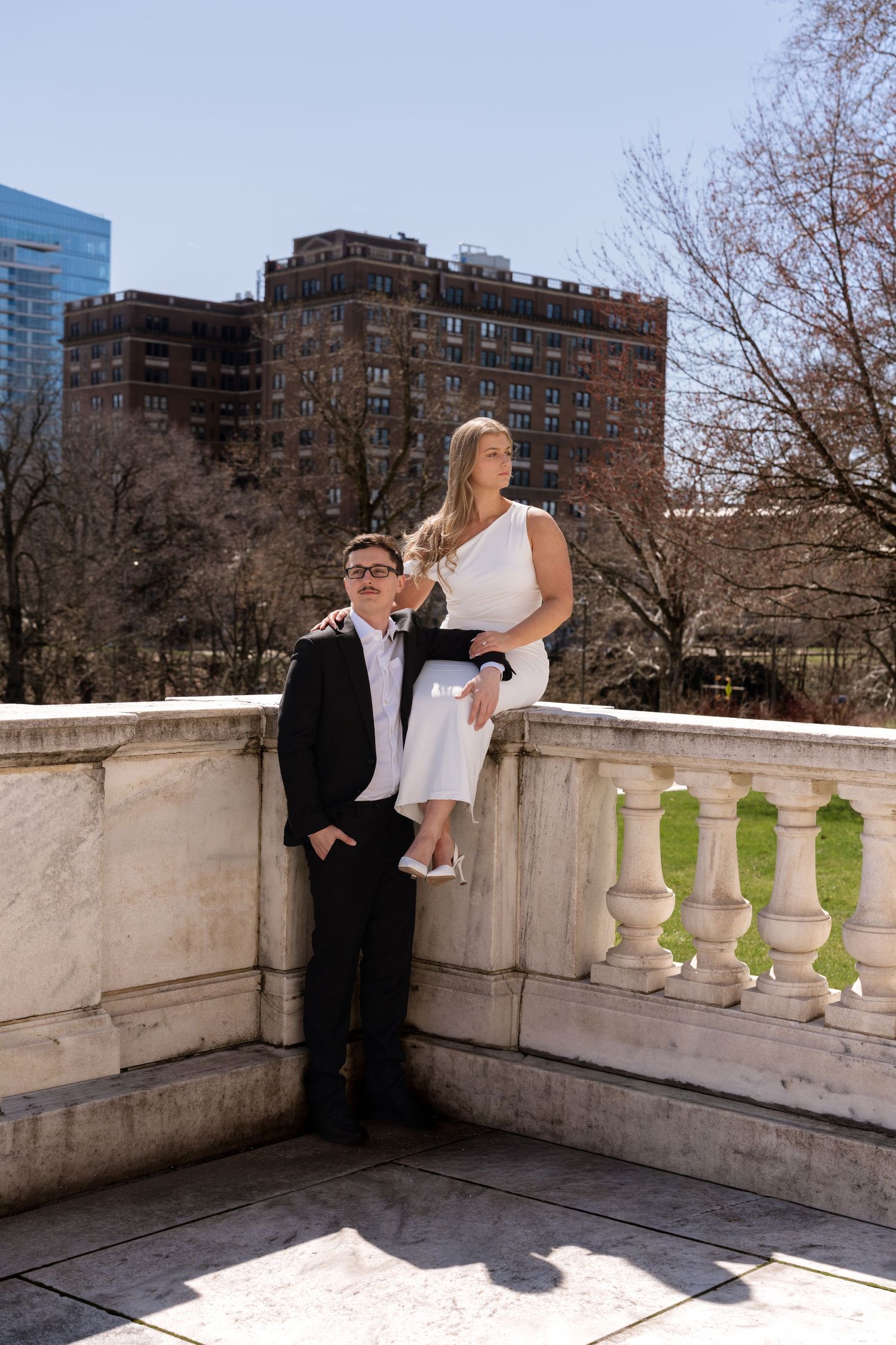 Couple in formal attire posing on a marble balcony in Boston Public Garden, woman in white dress seated on railing.