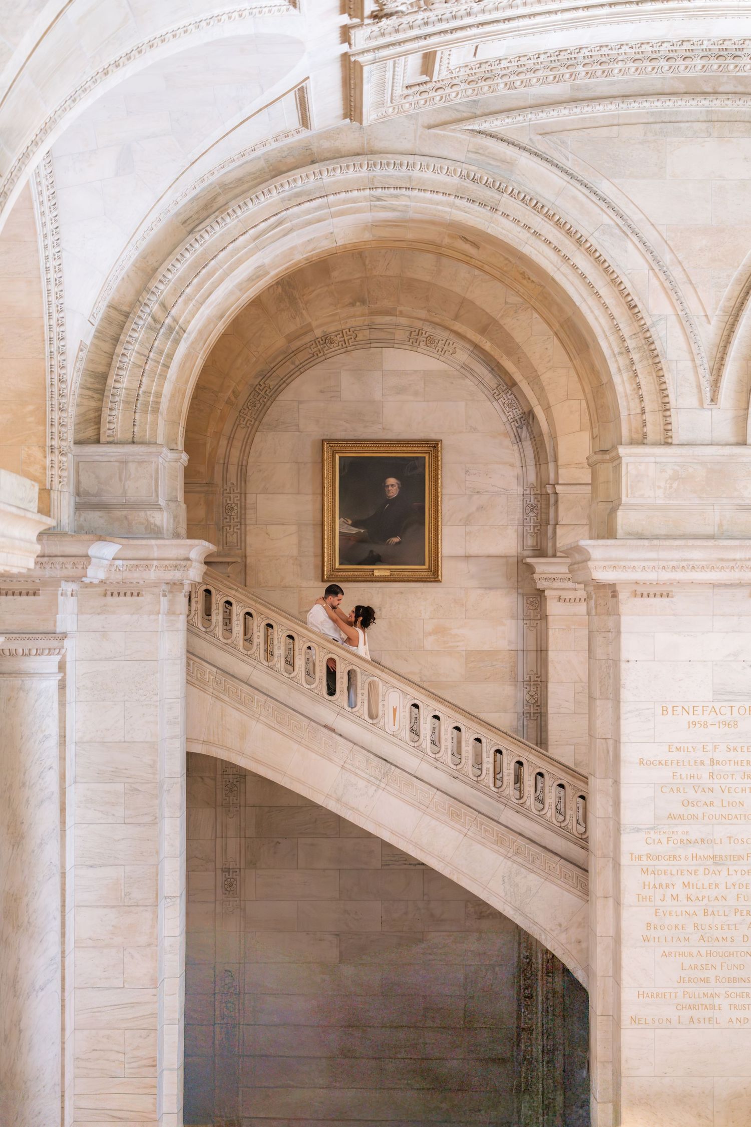 Couple sharing a romantic moment on grand marble staircase inside New York Public Library's elegant Beaux-Arts architecture.