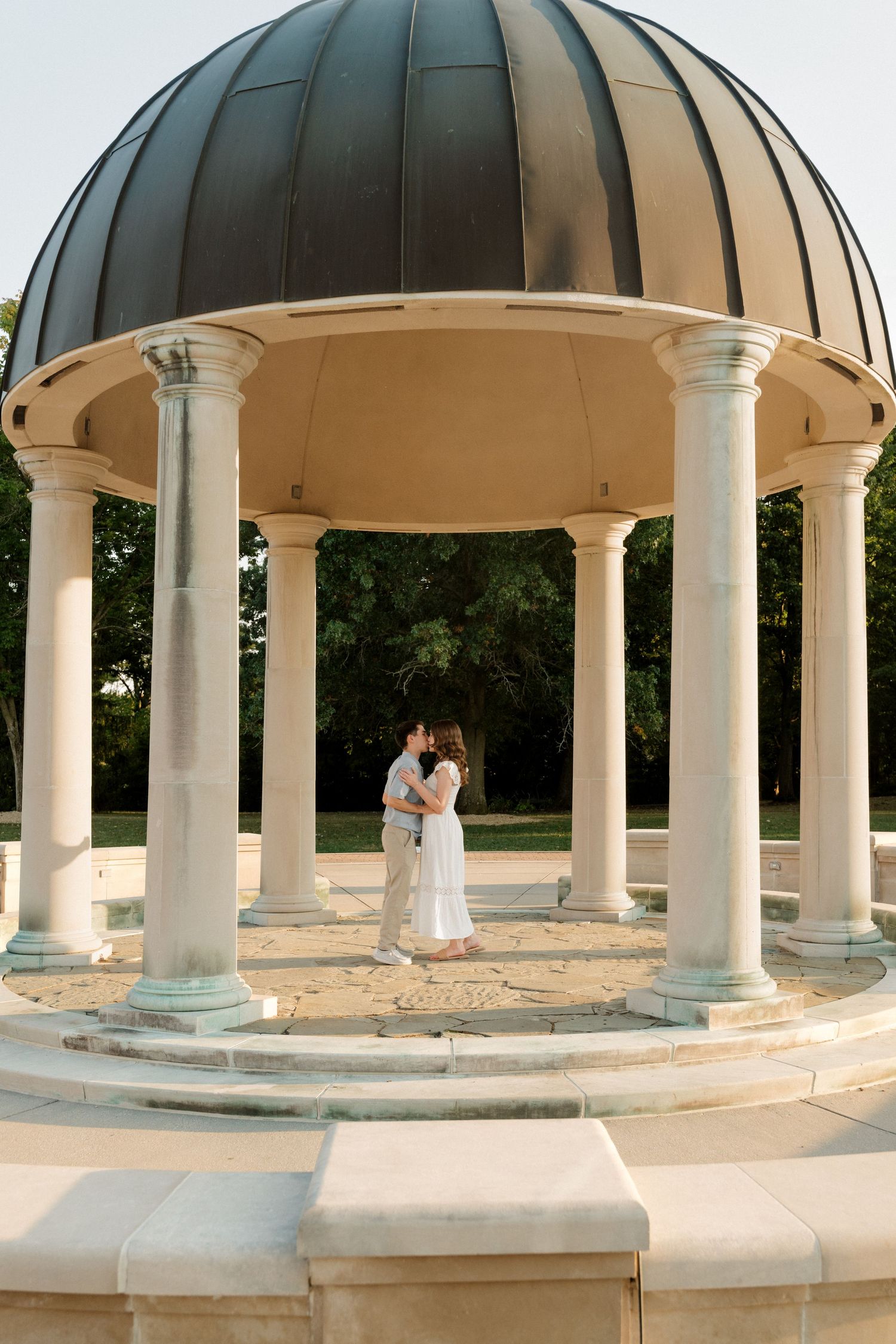 A romantic couple embraces under an elegant domed gazebo with white columns in a serene outdoor setting.