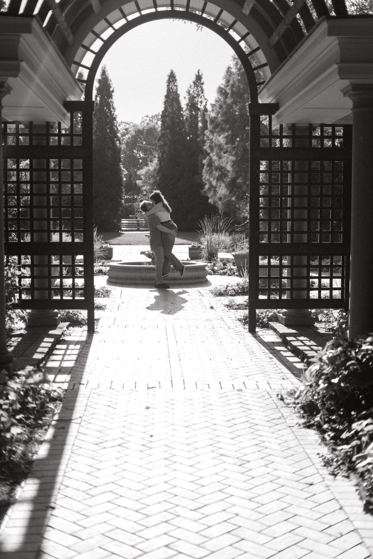 Black and white view through an arched garden trellis showing a brick pathway leading to a decorative fountain.
