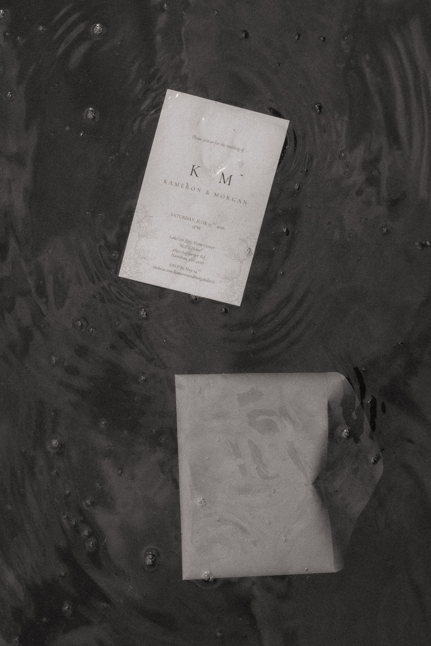 Black and white photo of a wedding invitation card and envelope floating in rippling water.