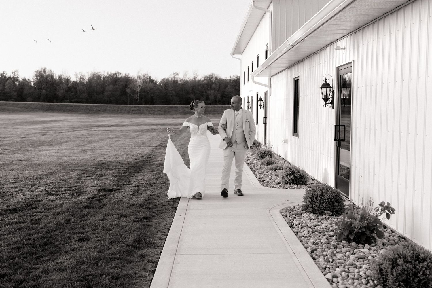 Black and white photo of a bride and groom walking hand in hand along a sidewalk beside a white barn venue.