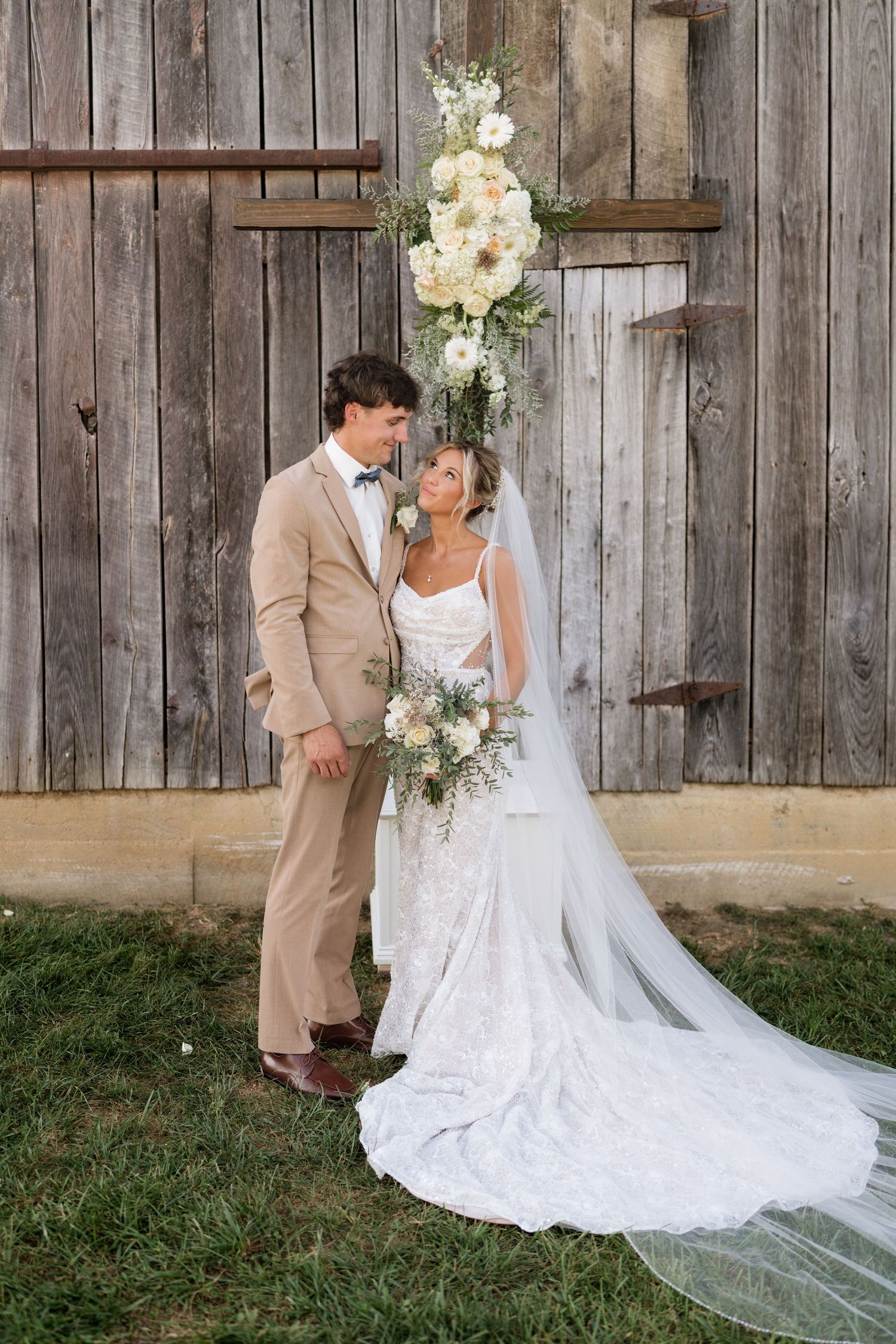 Bride and groom pose before rustic barn doors, she in lace gown with veil, he in tan suit with bow tie.