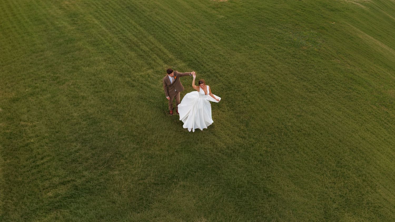 Aerial drone view of bride and groom holding hands walking through lush green field on wedding day.