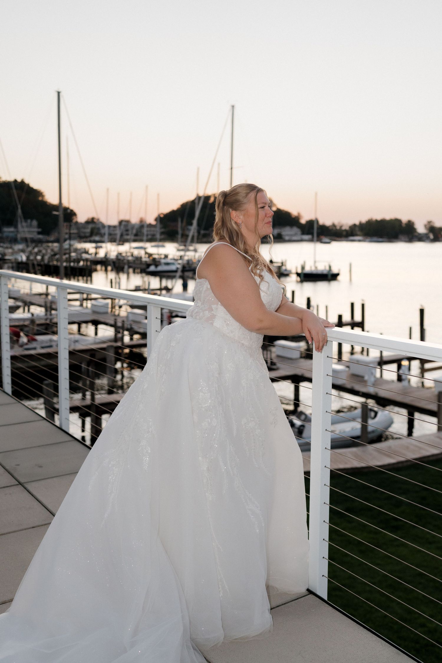 Bride in flowing white gown posing along marina walkway at sunset.