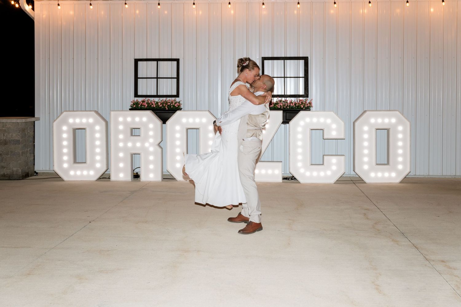 Couple shares romantic first dance lift in front of illuminated OROZCO marquee letters at rustic barn wedding reception.