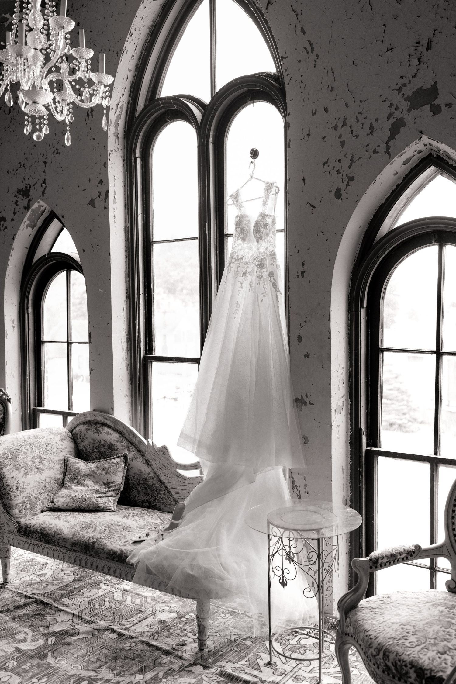 Black and white photo of an elegant wedding dress hanging in a vintage room with Gothic arched windows and a chandelier.