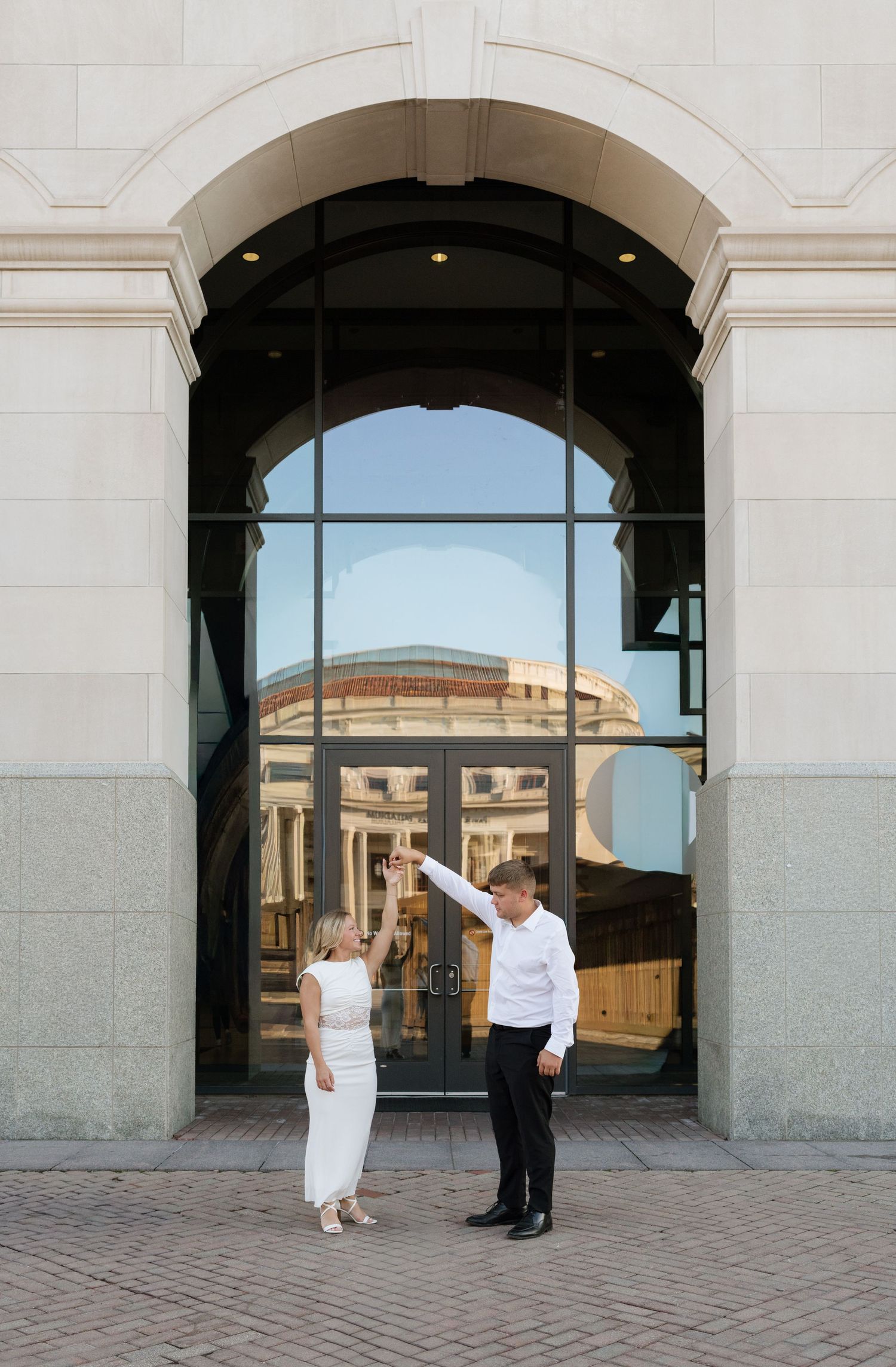 Couple dancing under an elegant stone archway with large glass doors reflecting a historic building on a sunny day.