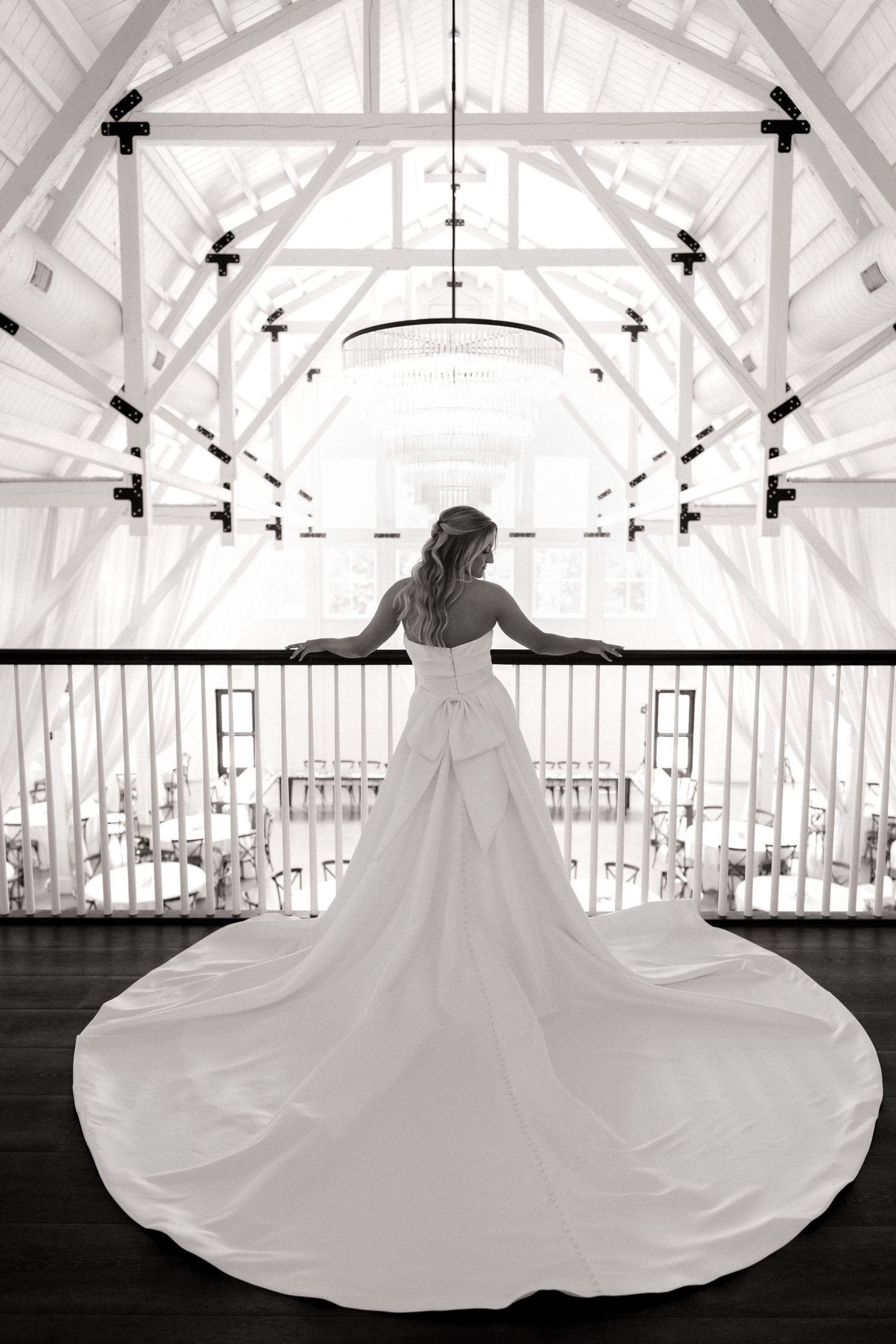 Black and white photo of a bride in a flowing white gown with long train standing on a balcony in an elegant venue.