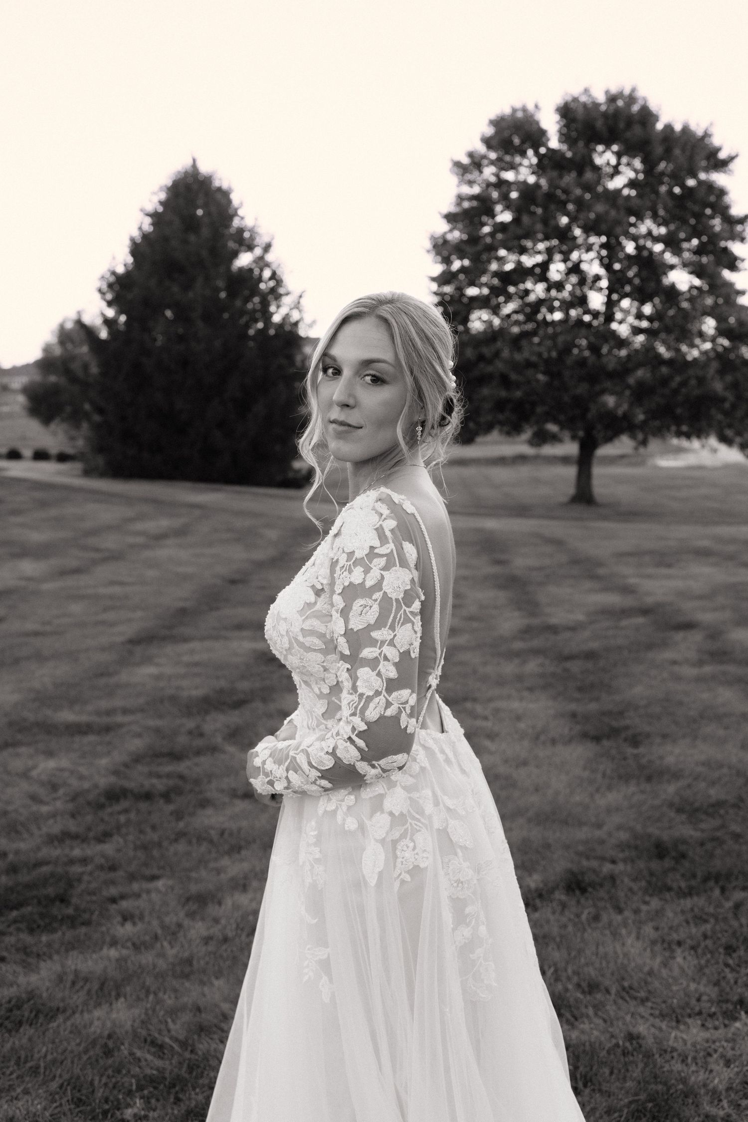 Black and white photo of a bride in a lace long-sleeve wedding gown posing outdoors on a grassy lawn.