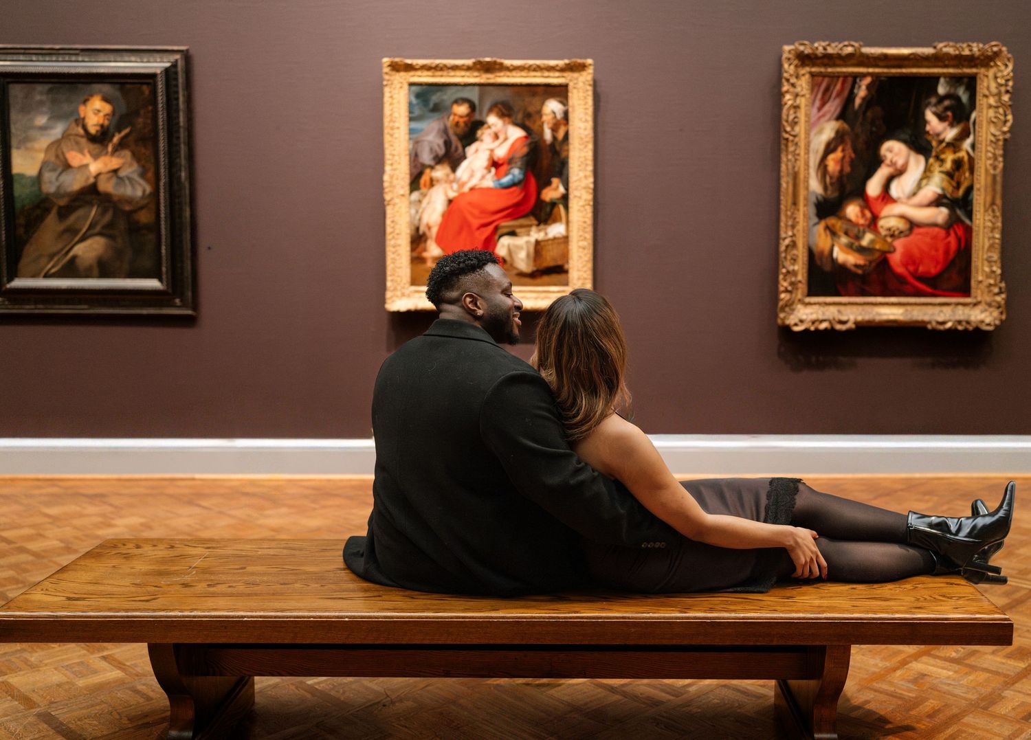 Museum visitors sit on wooden benches while viewing classical paintings in an art gallery.