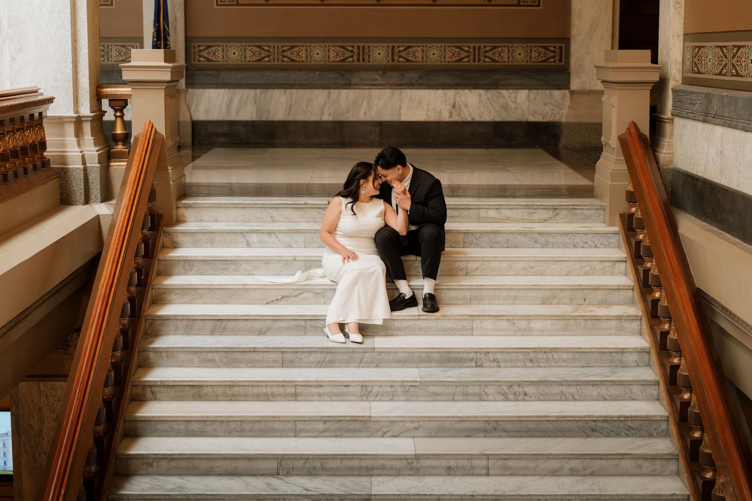 Bride and groom sit intimately on marble staircase in elegant venue, sharing a tender moment in wedding attire.