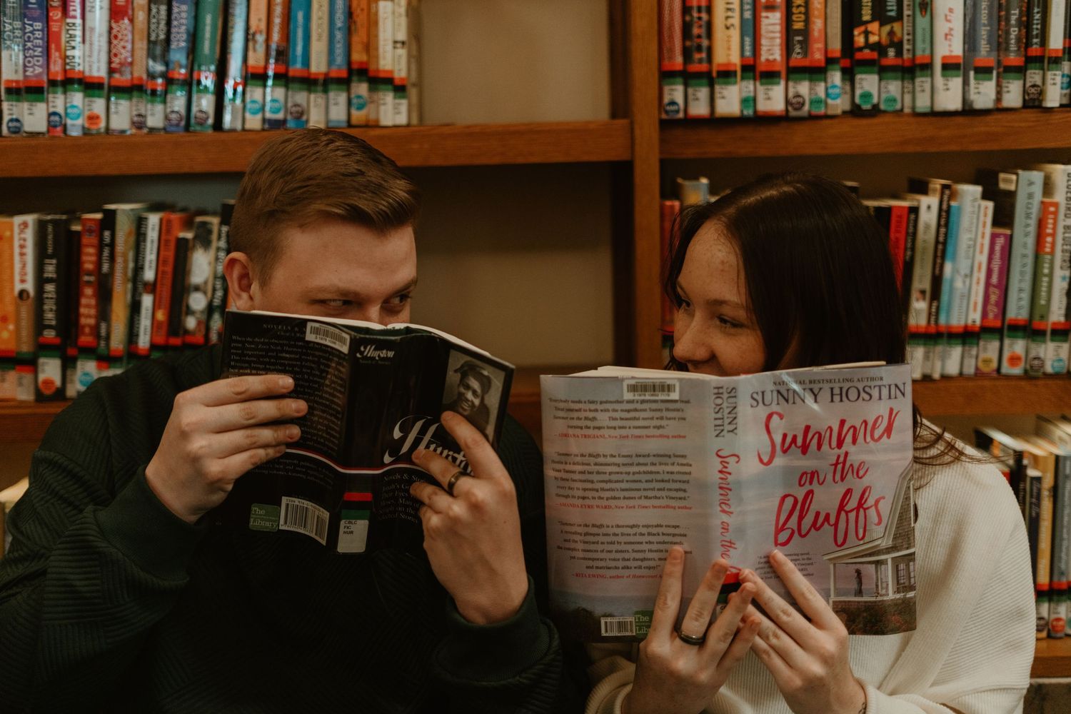 Person reading a book in a library with rows of bookshelves visible in the background.