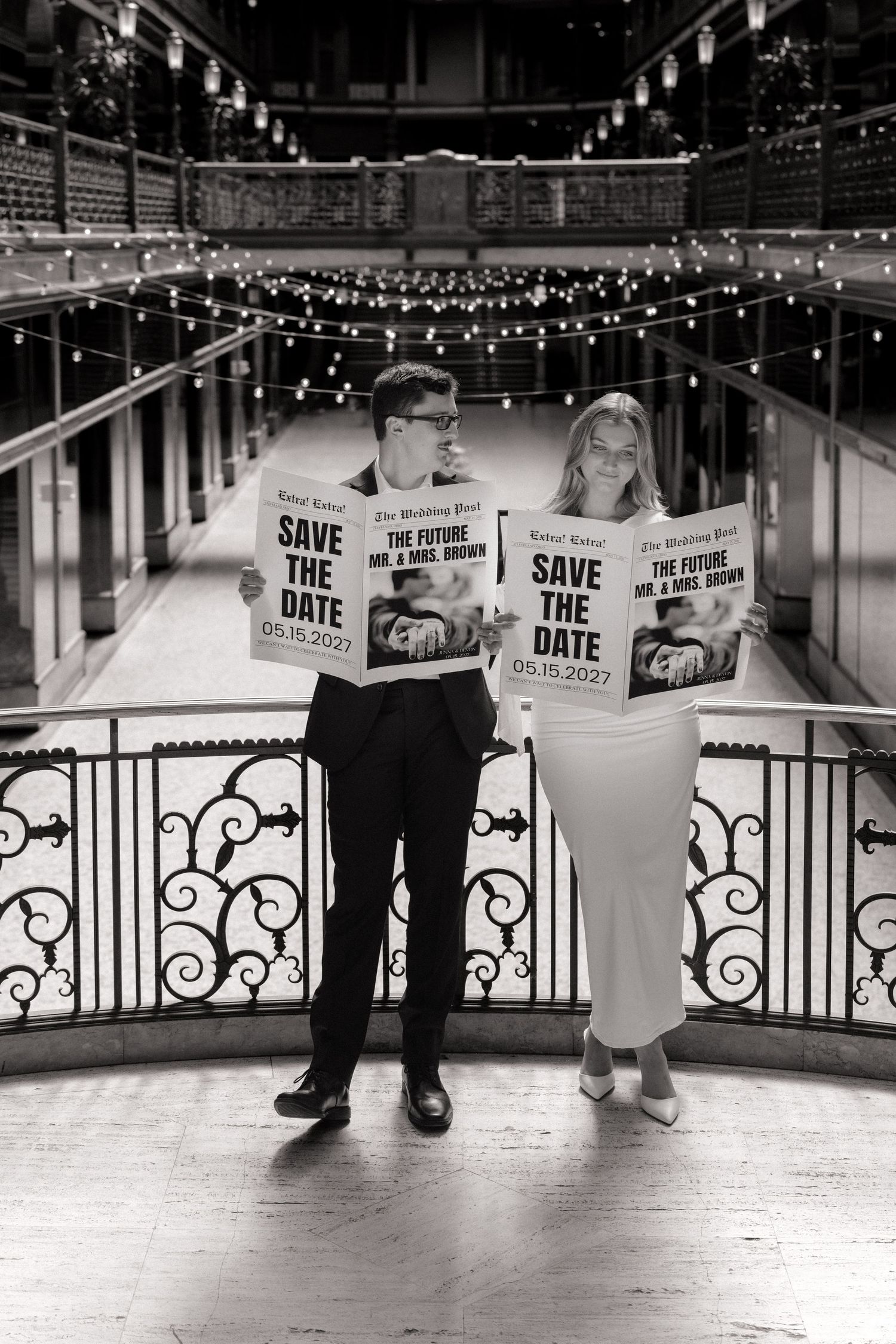 Black and white photo of couple holding Save the Date newspapers at elegant venue with string lights.