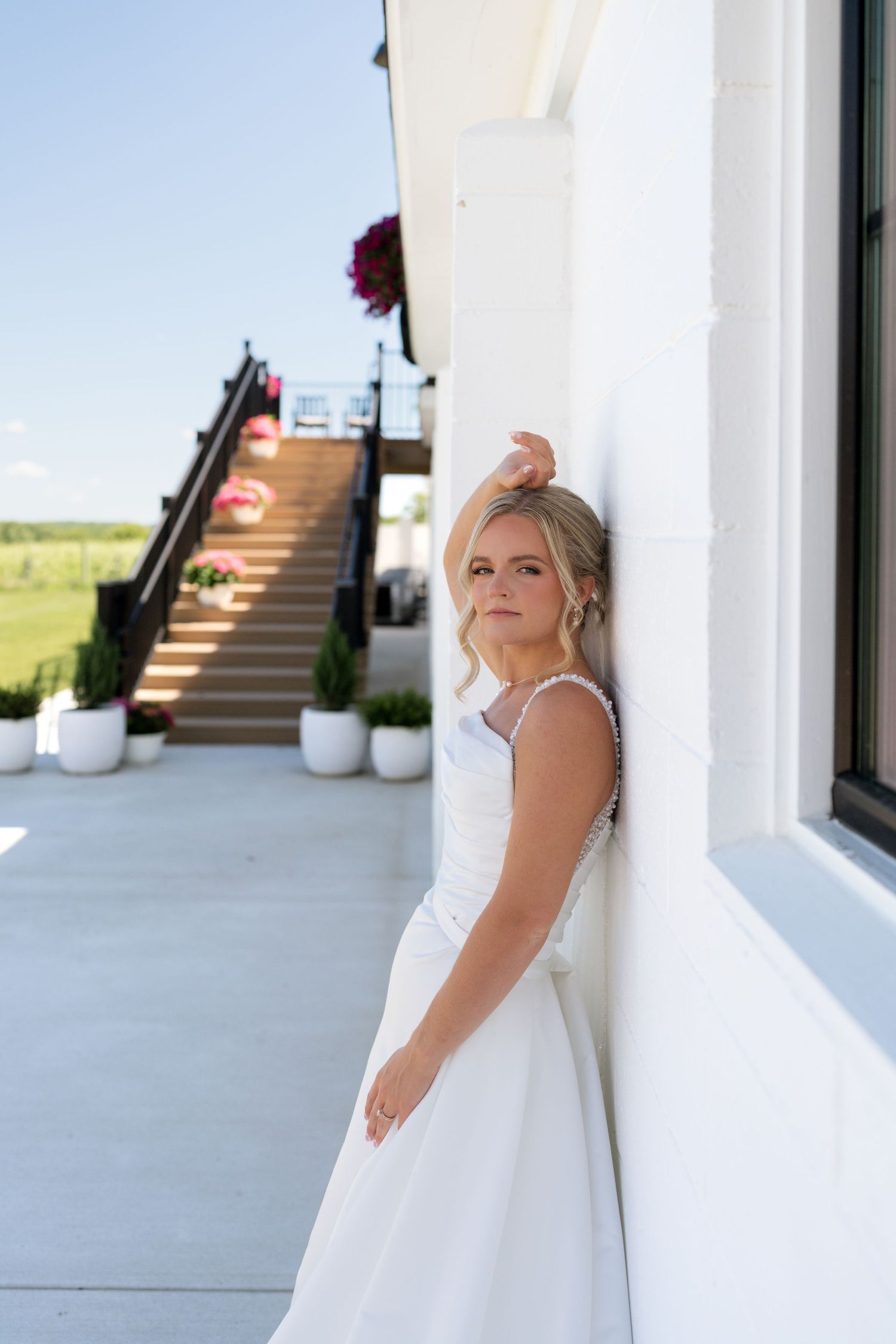 Elegant bride in white gown leaning against white wall at outdoor wedding venue on sunny day.