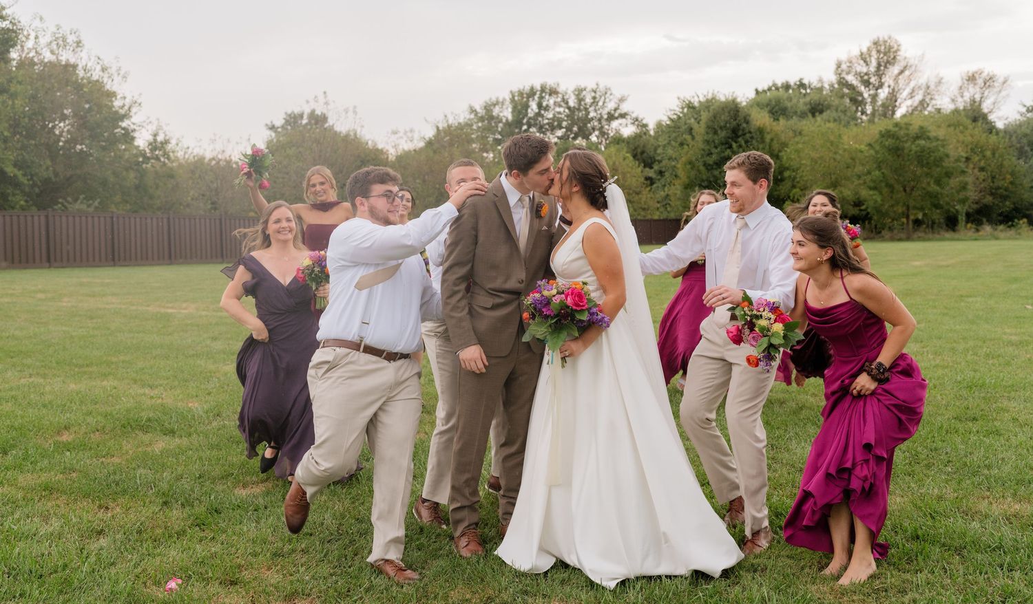 Wedding party playfully surrounds kissing bride and groom on green lawn, bridesmaids in purple dresses.