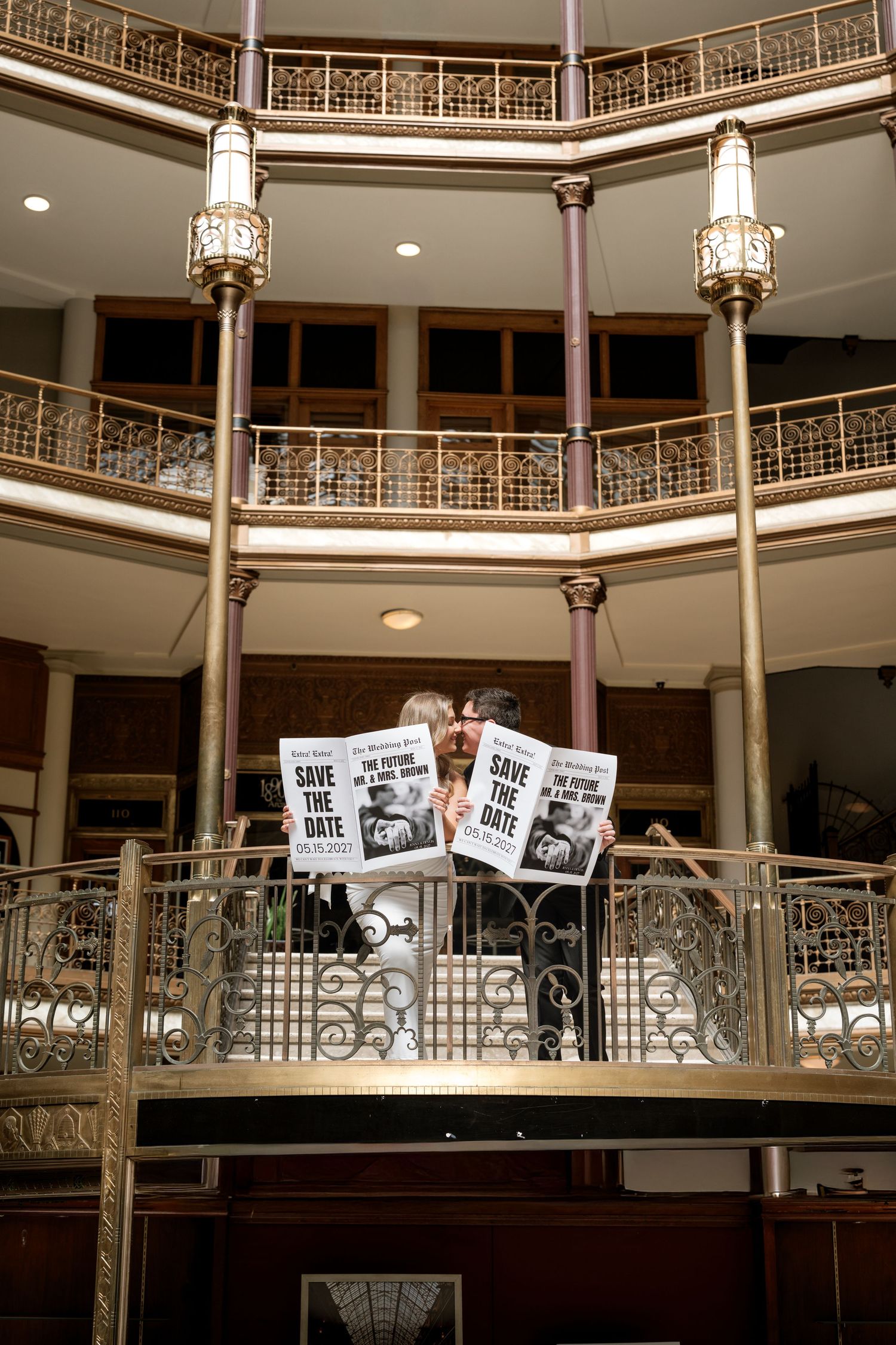 Couple holding Save the Date signs on ornate balcony in elegant multi-level venue with decorative railings and gold lamp posts.
