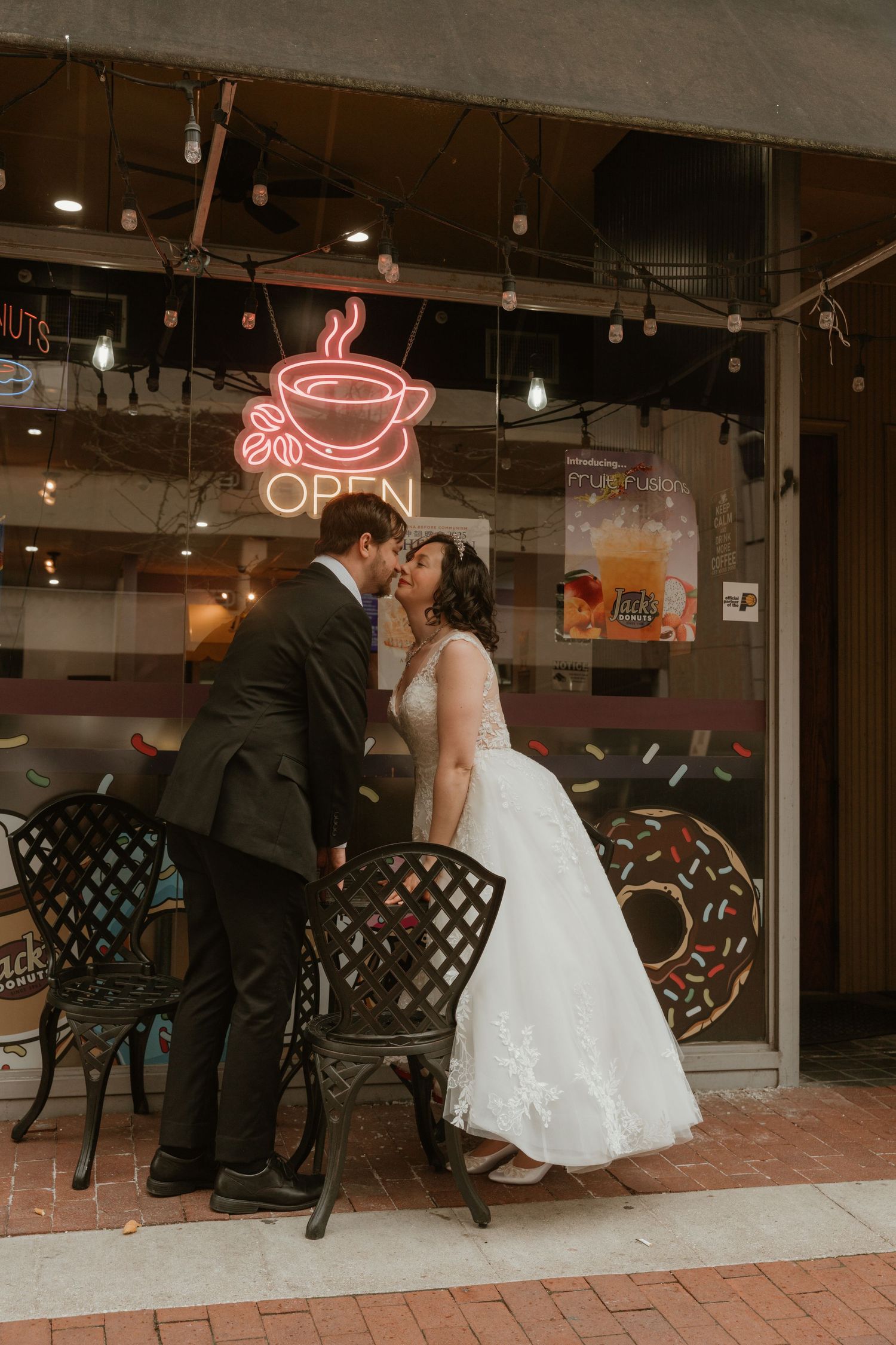 A romantic wedding couple shares a kiss outside a cafe with a glowing neon coffee cup sign in the window.