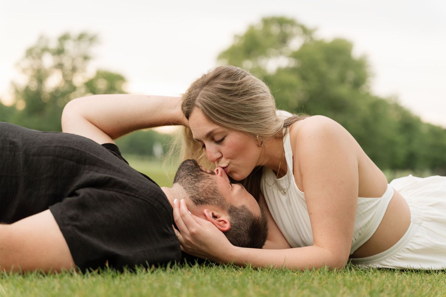 Couple lying on green grass in a romantic outdoor setting, sharing a tender kiss during a sunny engagement photo session.