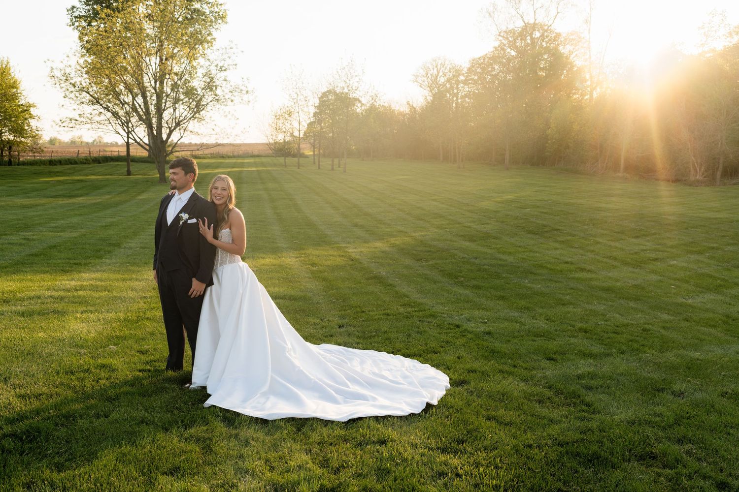 Bride and groom posing together at sunset on a green lawn, brides long white train flowing gracefully behind her.