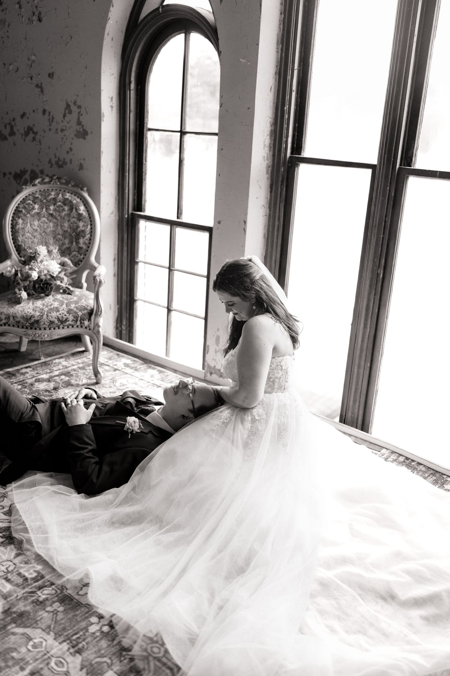 Black and white wedding photo of bride in tulle gown sitting with groom lying on ornate rug by arched windows.
