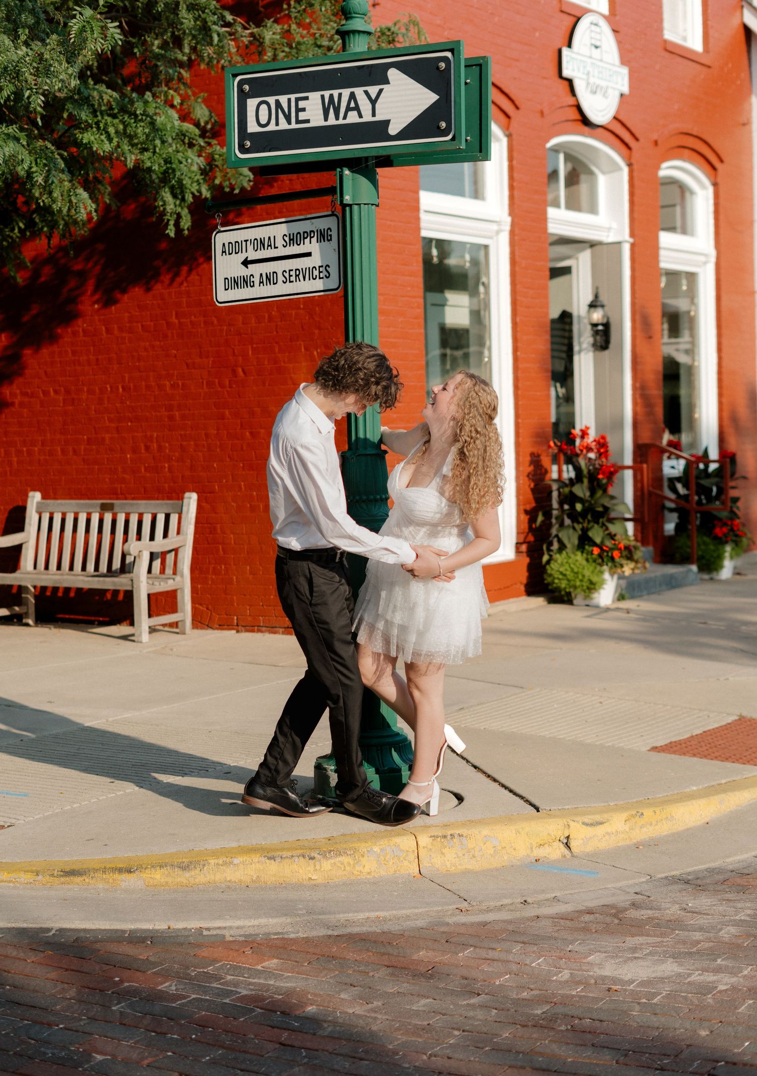 A couple in formal attire walks past a red brick building with a one-way street sign visible in the urban setting.