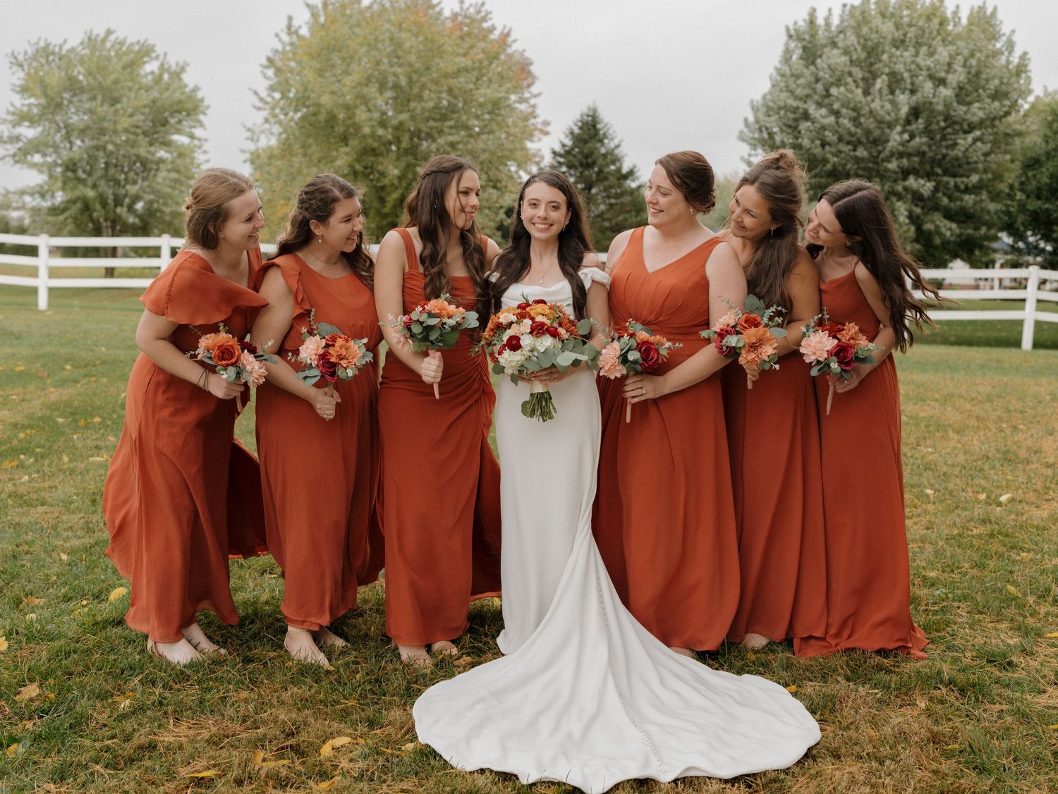 Bridesmaids in warm terracotta dresses gather around bride in white gown on fall grass.