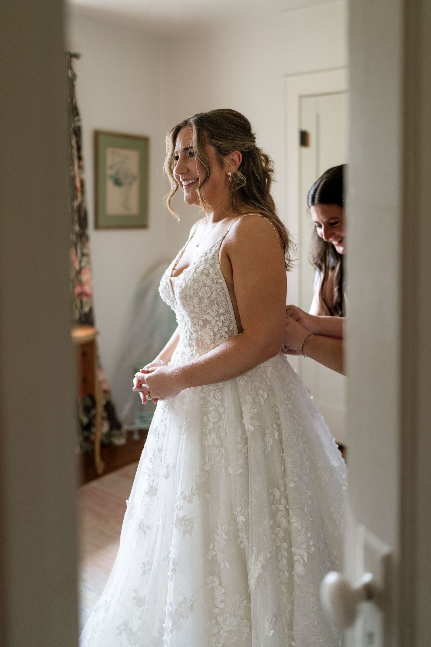 Smiling bride in floral lace ball gown being helped into her wedding dress by a bridesmaid.