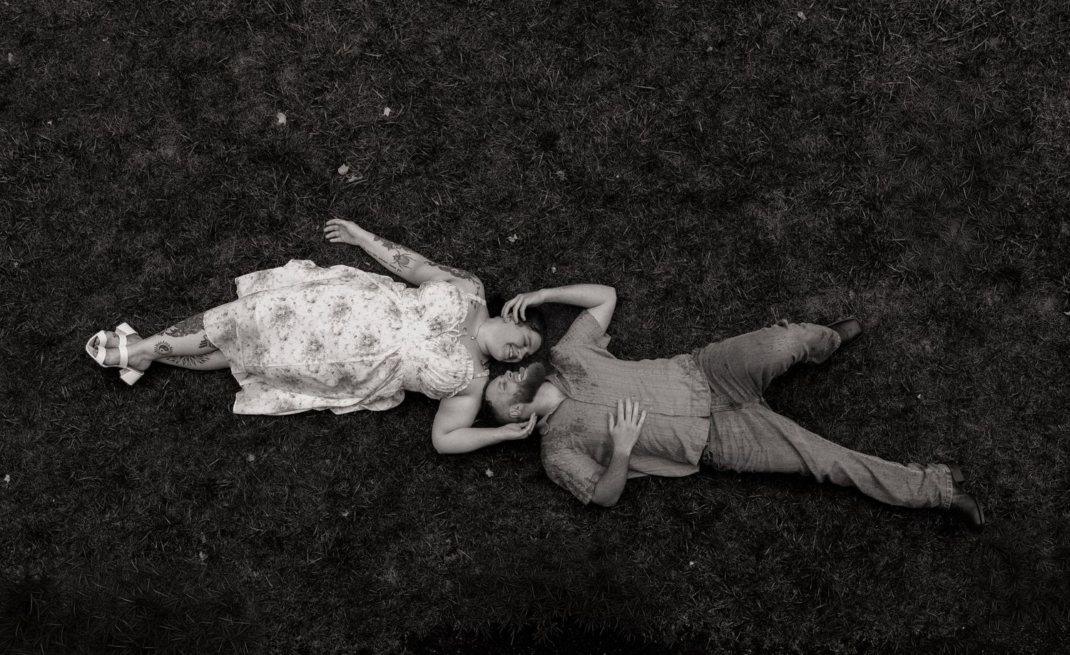 Black and white photo of a couple lying on grass, woman in floral dress and man in casual clothes, aerial view.